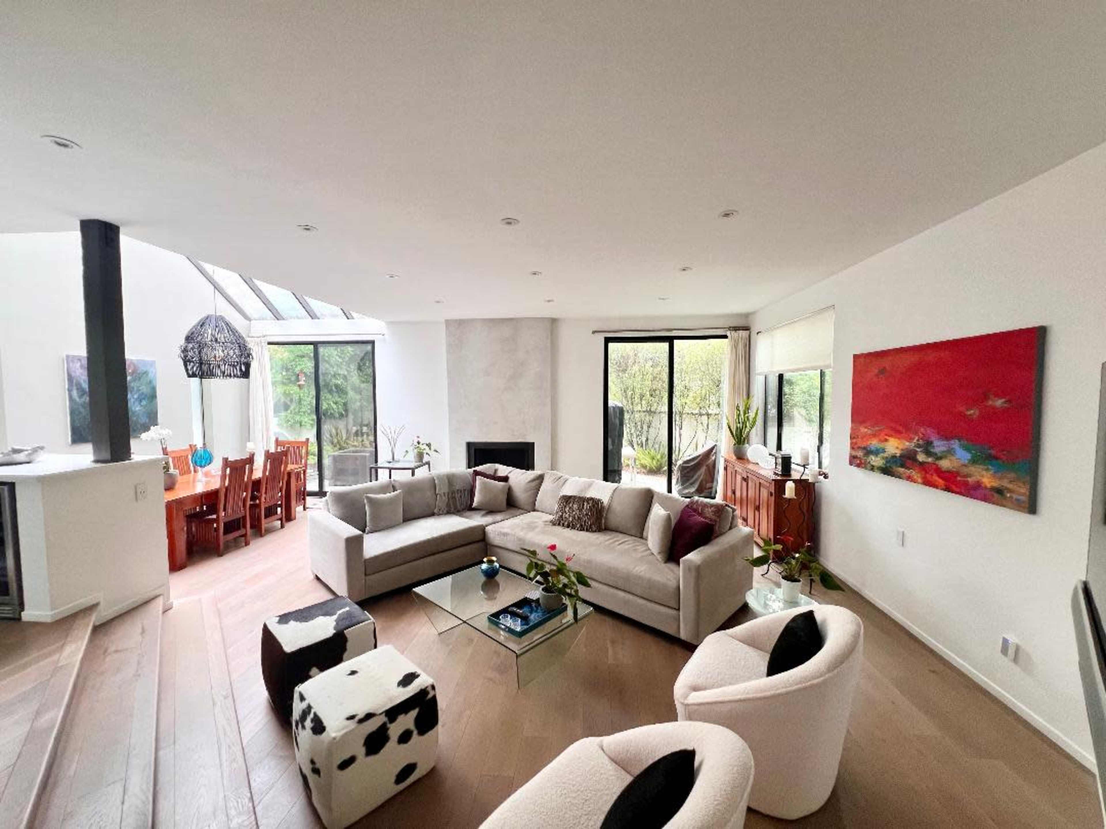 The image shows a modern living room with a sectional sofa, a coffee table, and large windows that provide natural light, leading to a dining area in the background.