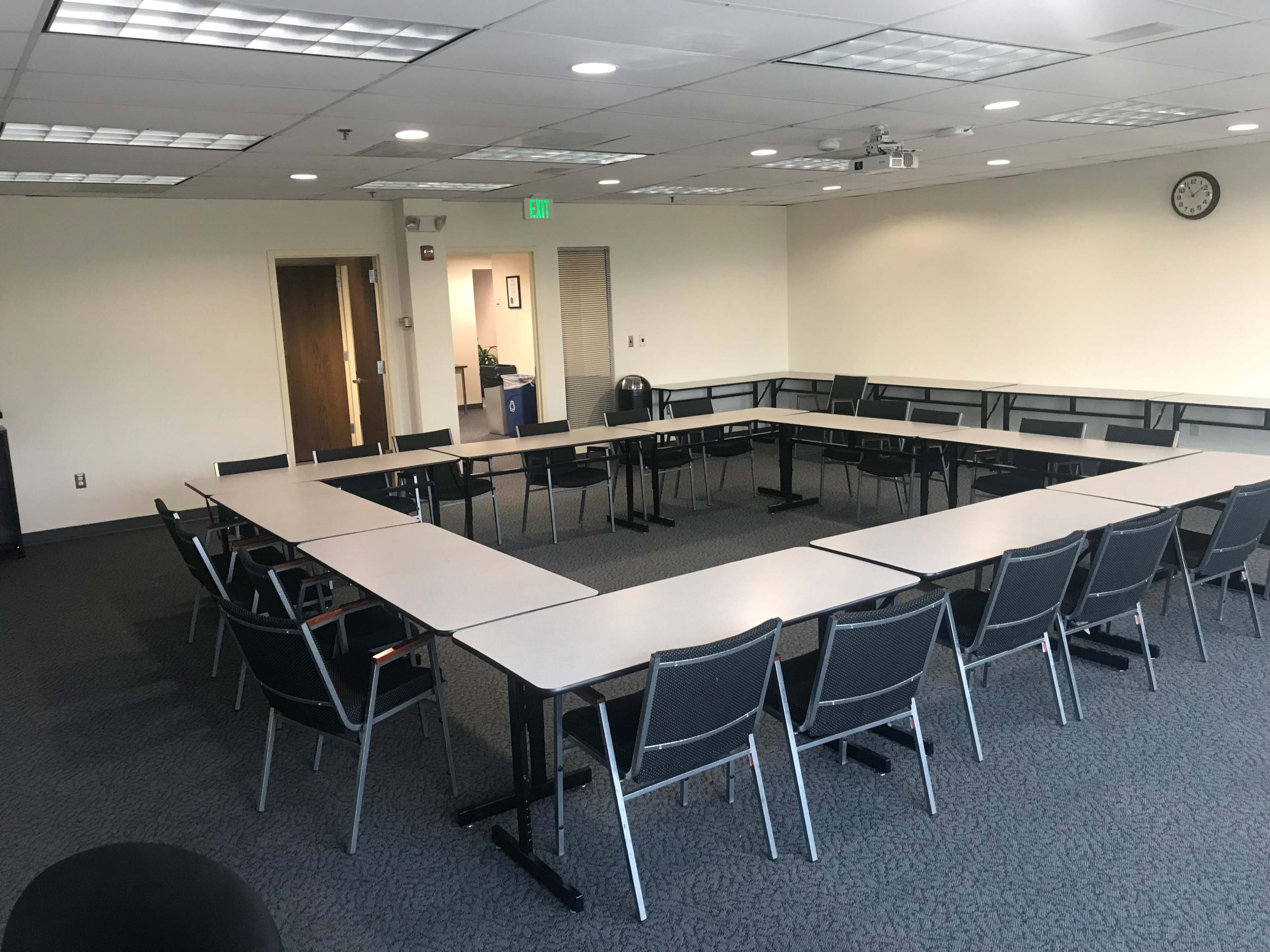A rectangular arrangement of tables and chairs is set up in a meeting room, with a clock and doors visible in the background.