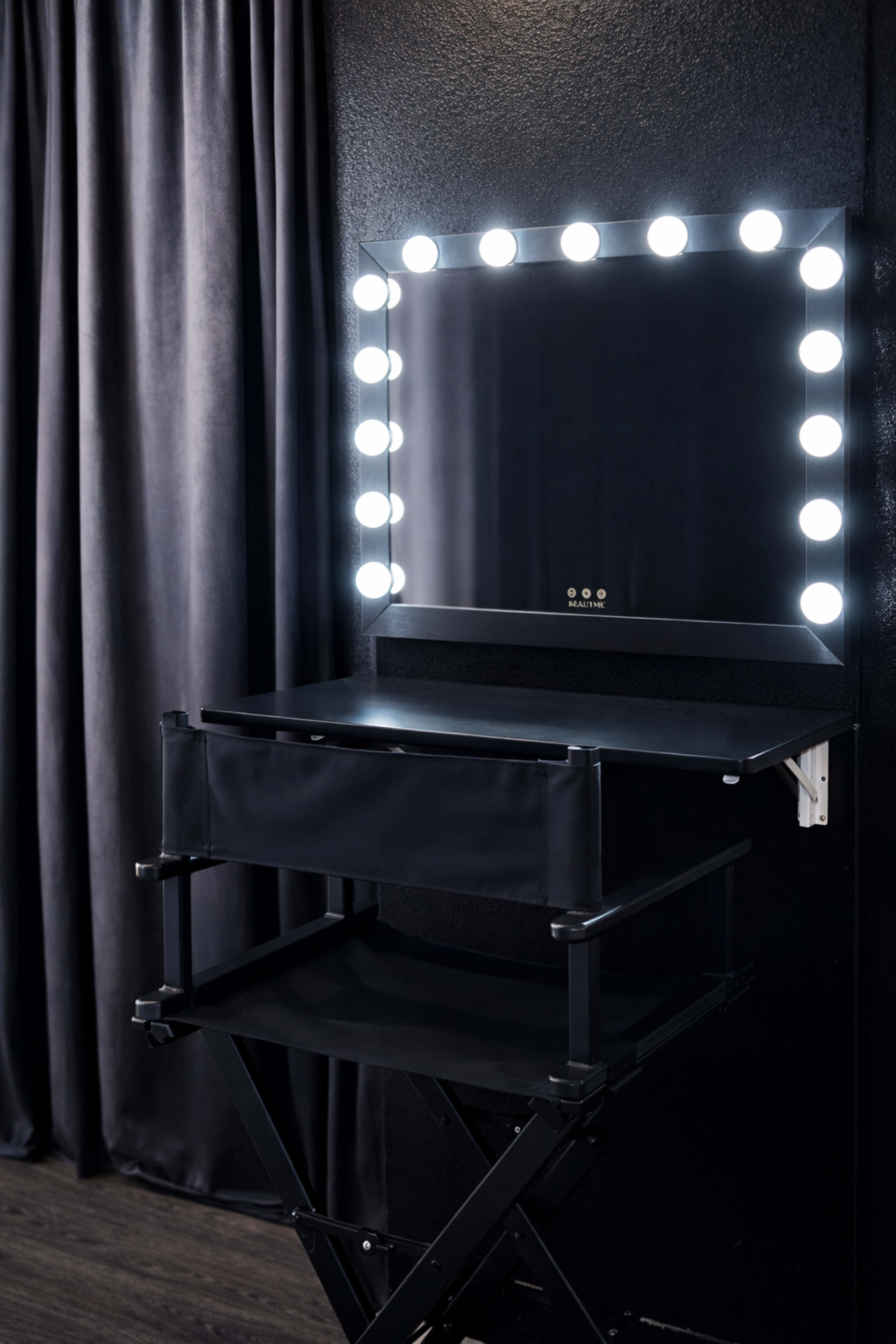 The image shows a black vanity mirror with lights mounted on the wall above a foldable director's chair in front of it.
