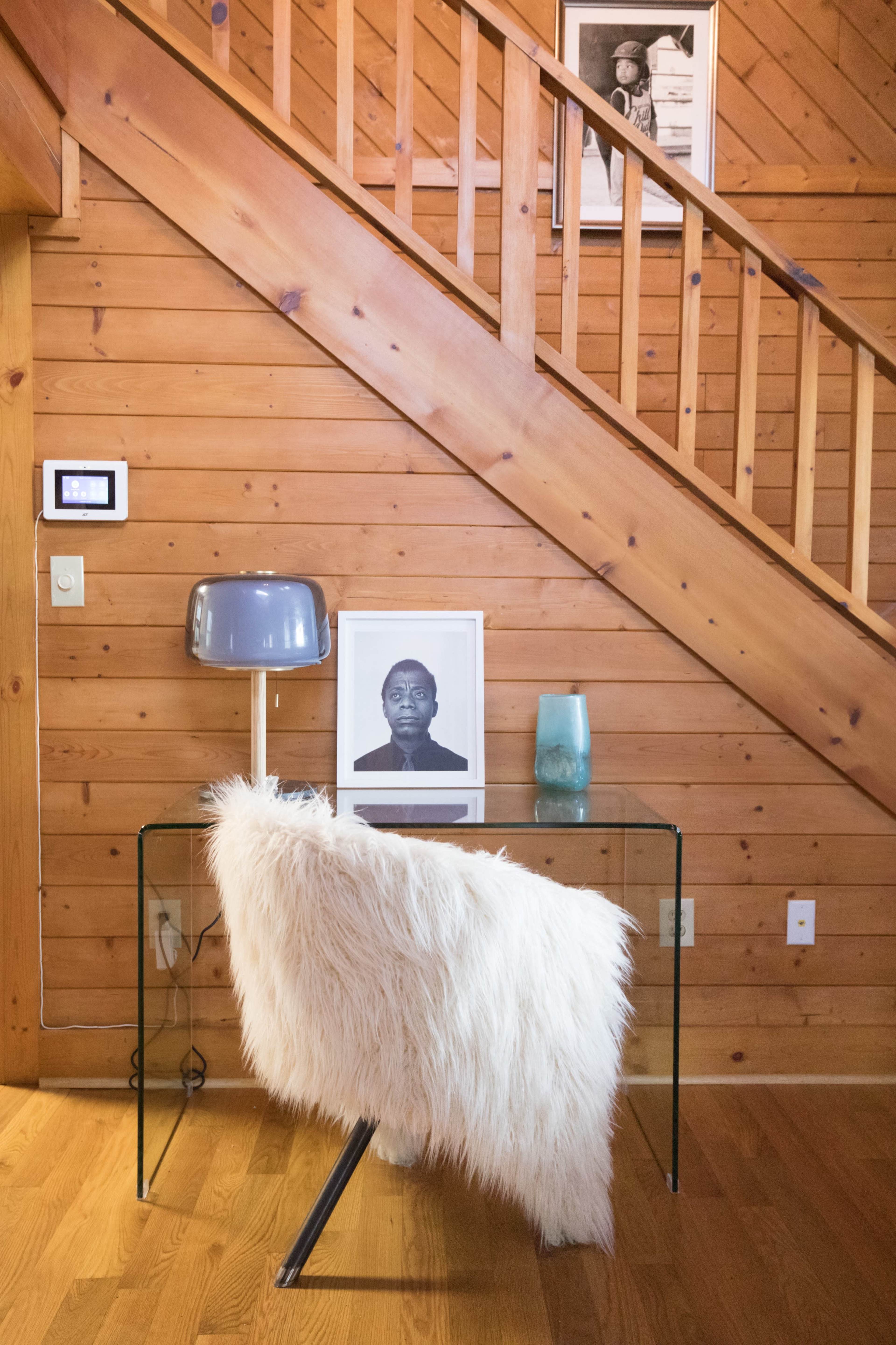 A clear glass desk with a fluffy white chair is positioned near a wooden staircase, accompanied by a lamp and a framed portrait on the wall.