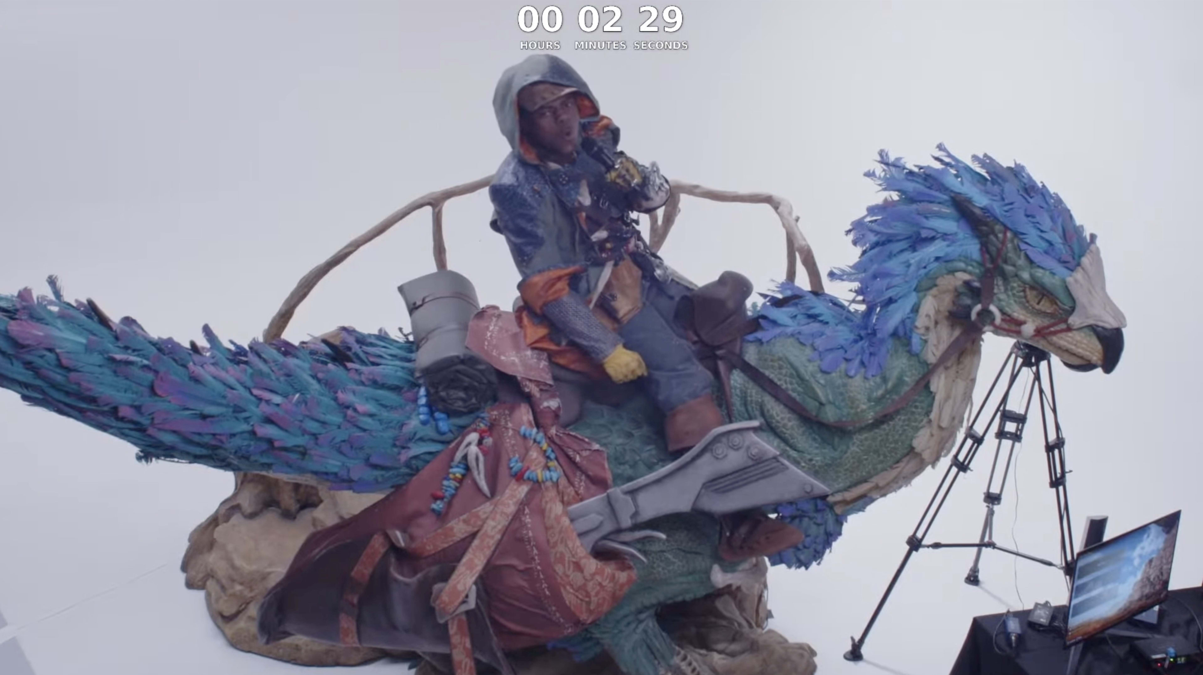 A person in a blue and brown outfit sits atop a large, vividly colored bird-like creature, surrounded by cameras and a timer displaying "00:02:29."