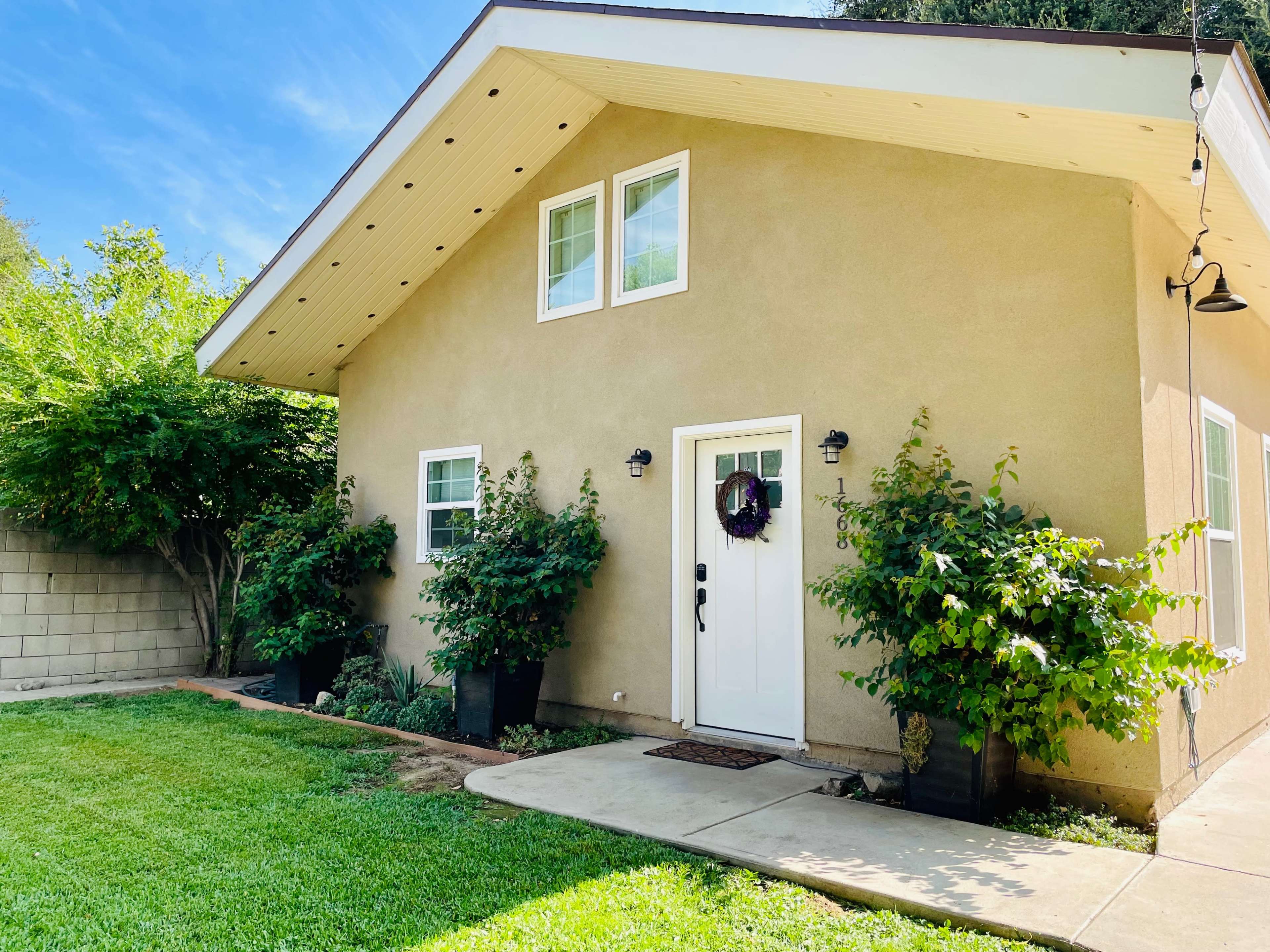A small, tan house with a peaked roof has white trim and is surrounded by greenery and potted plants.