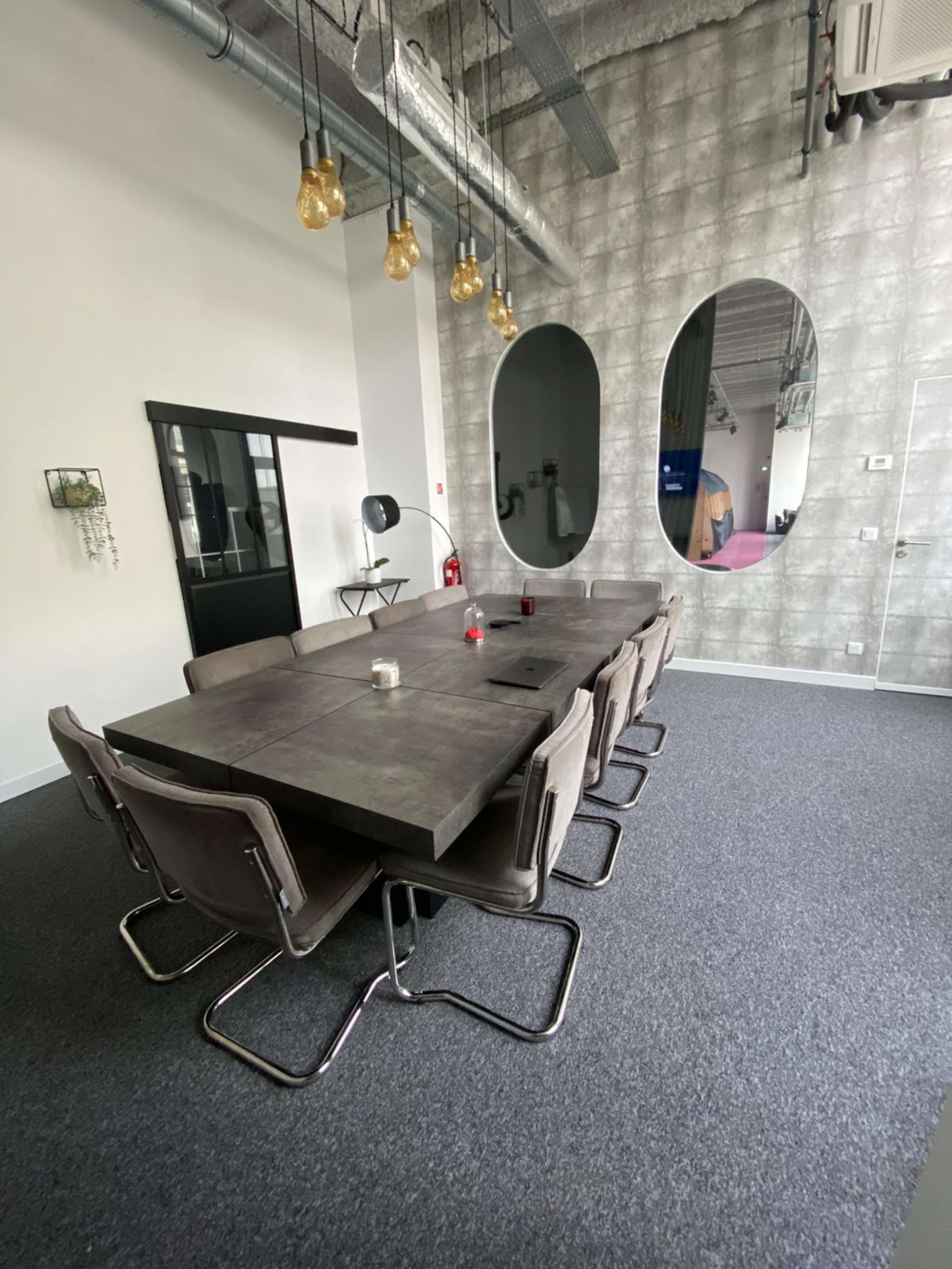 A long concrete conference table with eight chairs is set up in a modern meeting room that features two large oval mirrors and exposed ductwork on the ceiling.