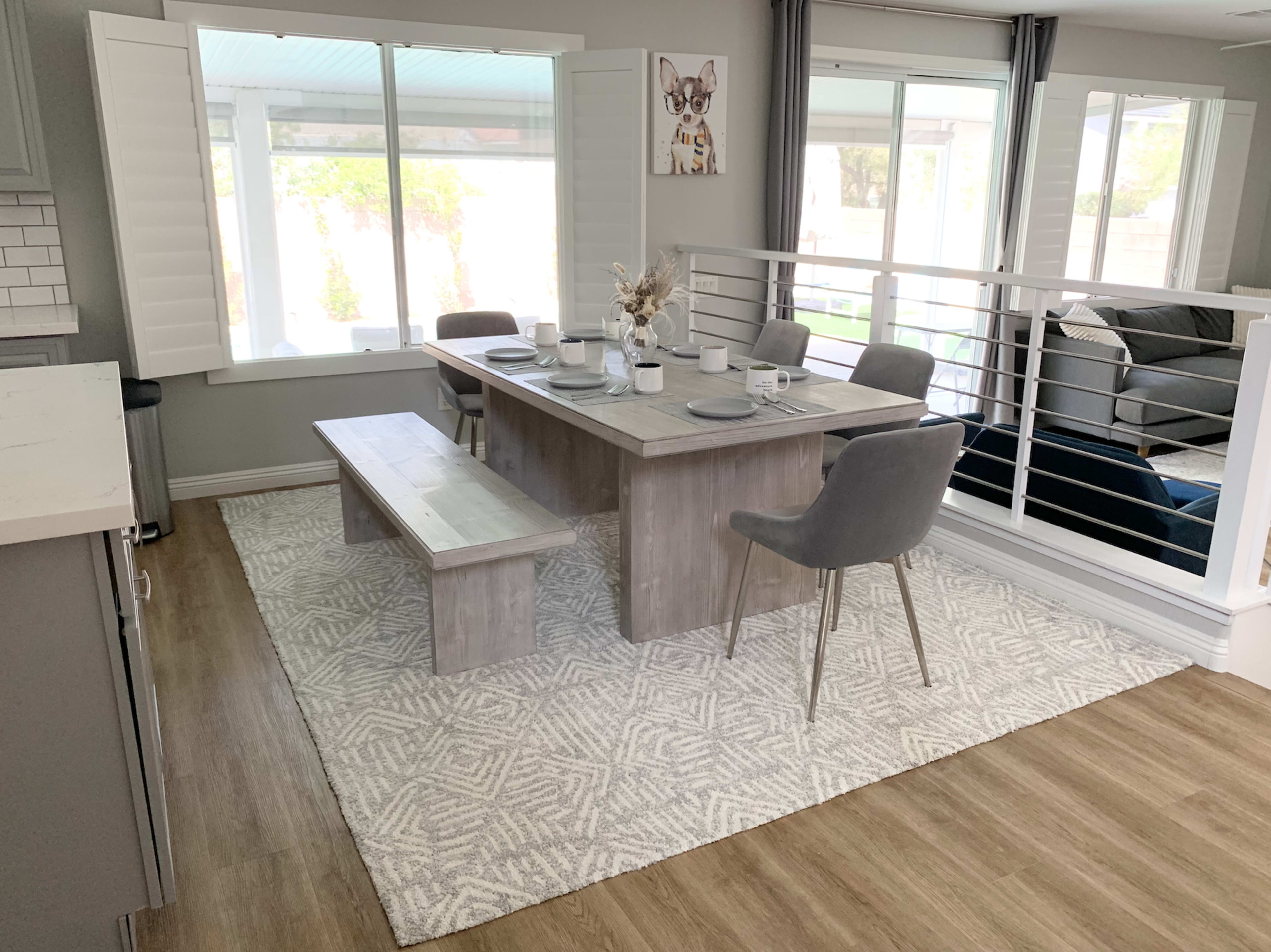 A modern dining area features a wooden table with gray chairs and a bench, set on a patterned rug under natural light from large windows.
