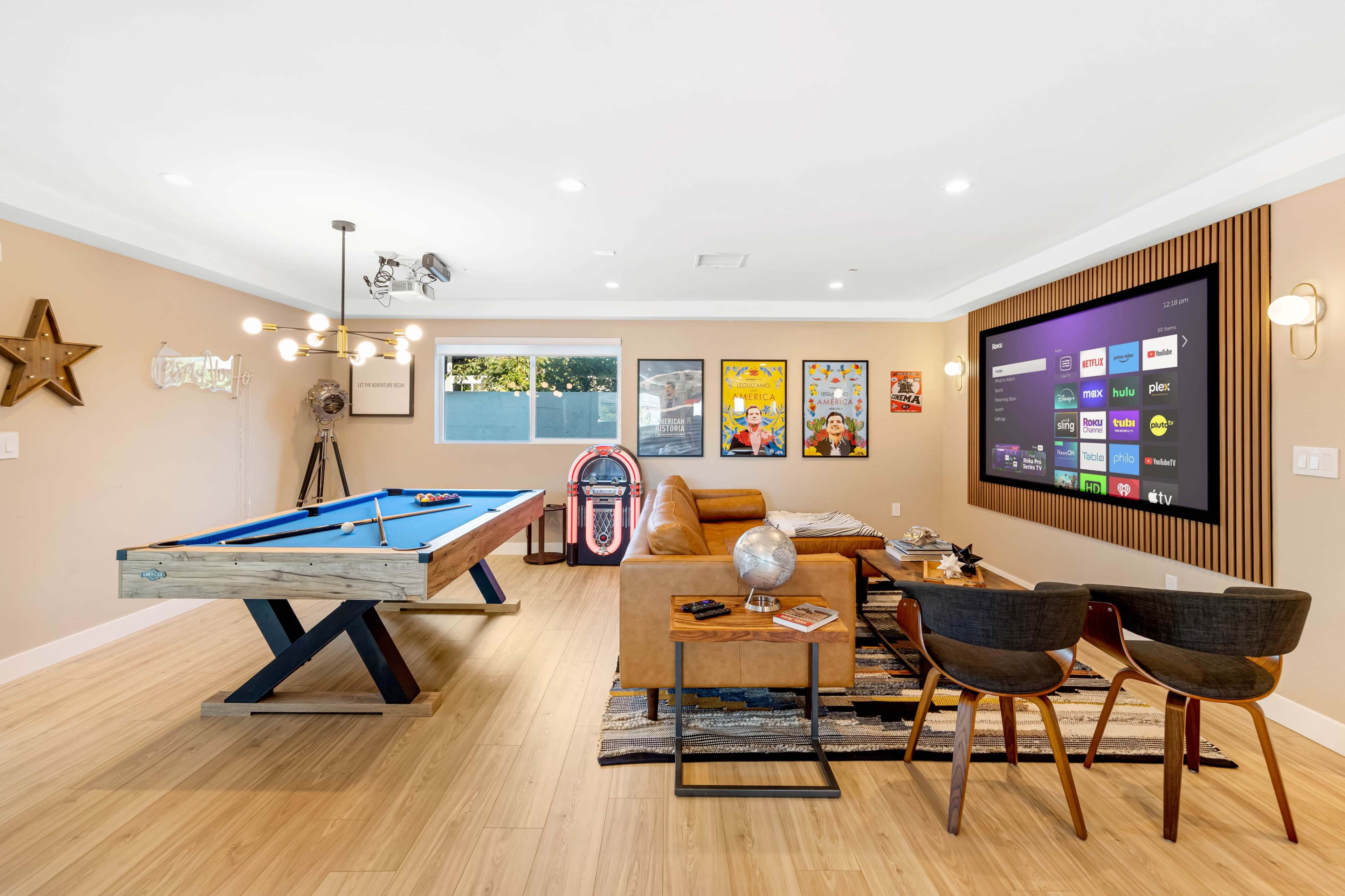 The image shows a modern game room featuring a blue pool table, a brown sofa, and a large wall-mounted television.