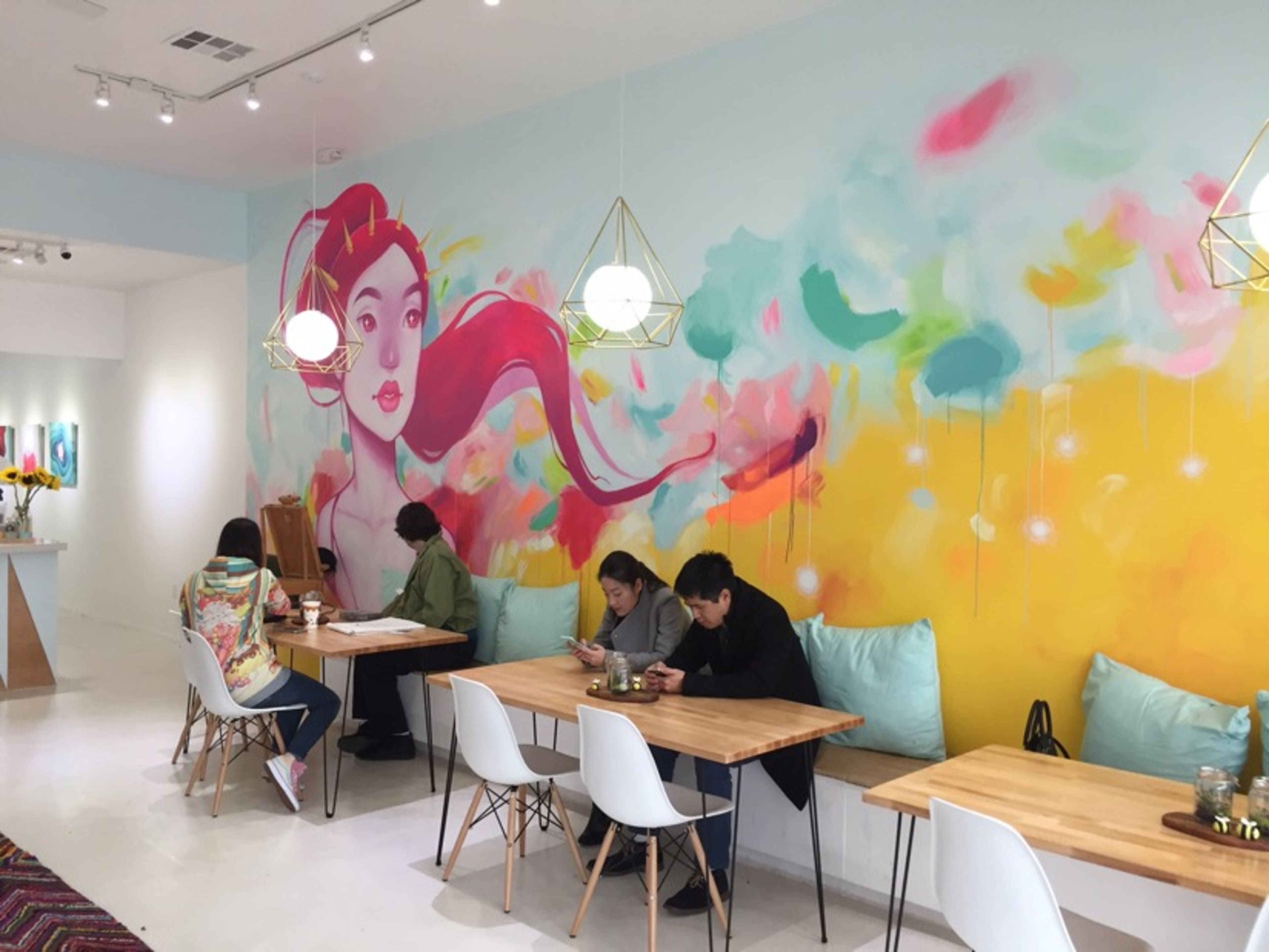 A colorful café interior with a large mural of a woman on the wall and several patrons seated at wooden tables.