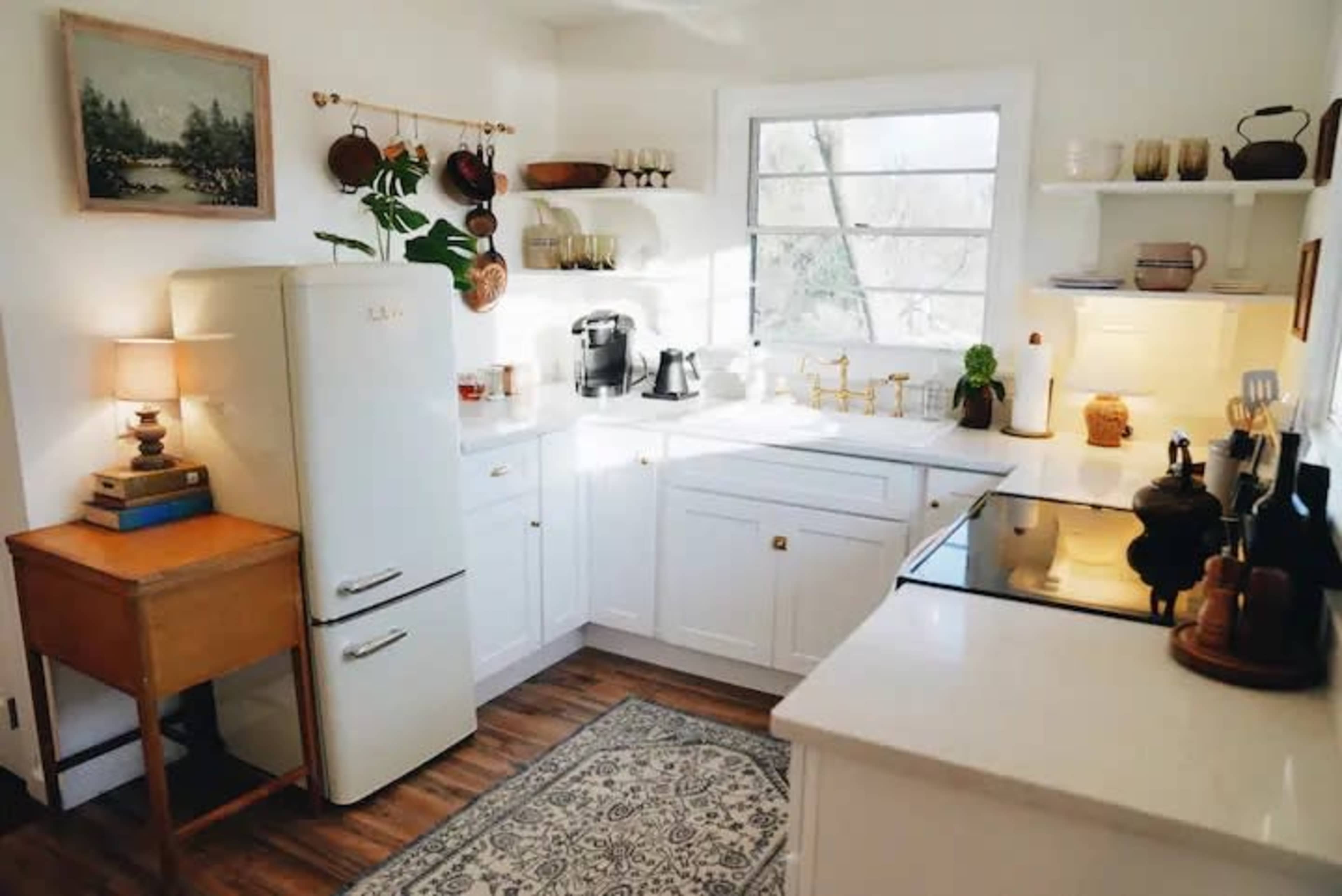 A bright kitchen features a retro refrigerator, wooden cabinetry, a small table with a lamp, and various kitchen appliances on the counters.