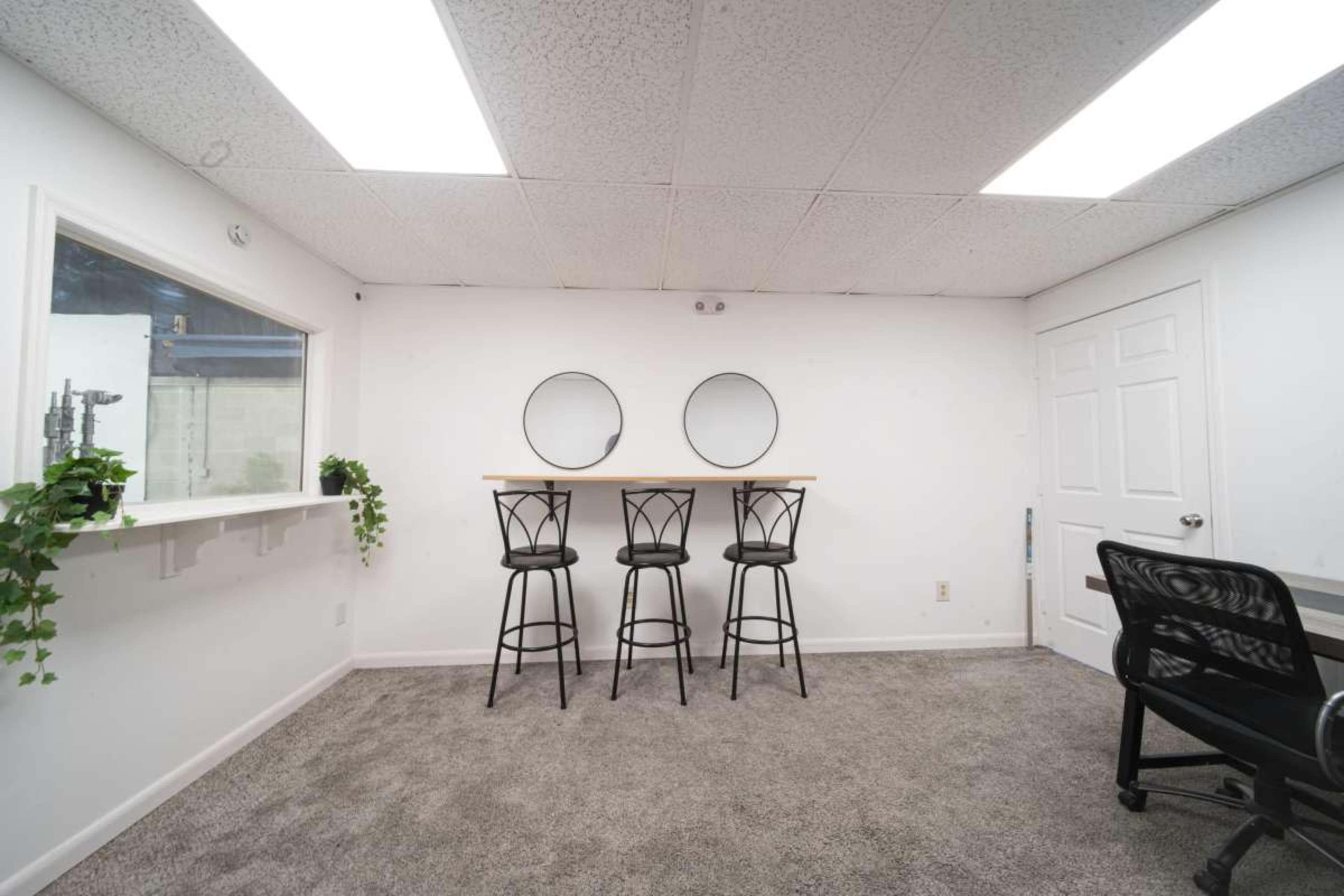 The image shows a minimalistic room featuring three high stools at a bar-height table, two circular mirrors on the wall, and a desk with a black office chair.