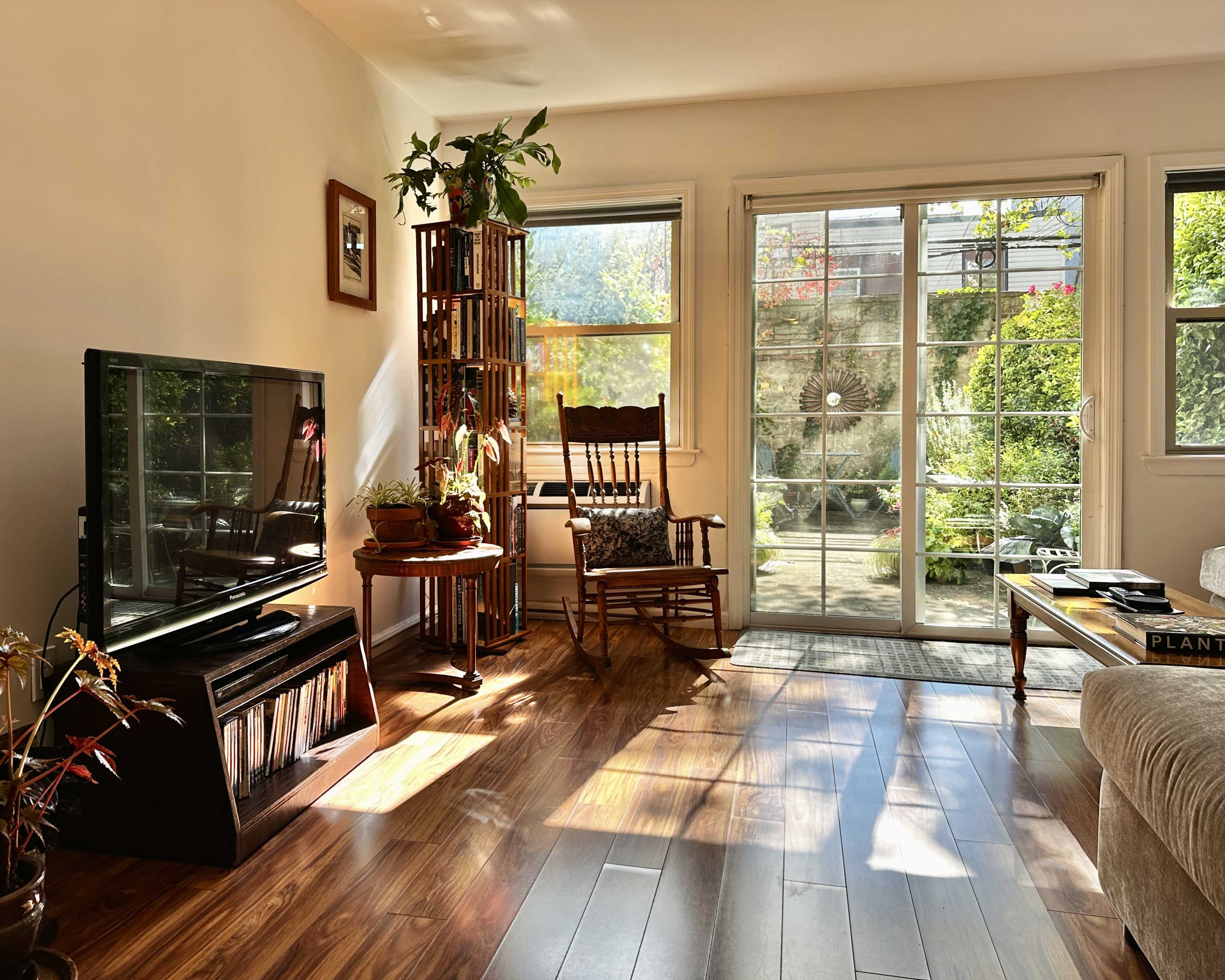 A sunlit living room features a rocking chair, a television, and a view of a landscaped garden through glass doors.