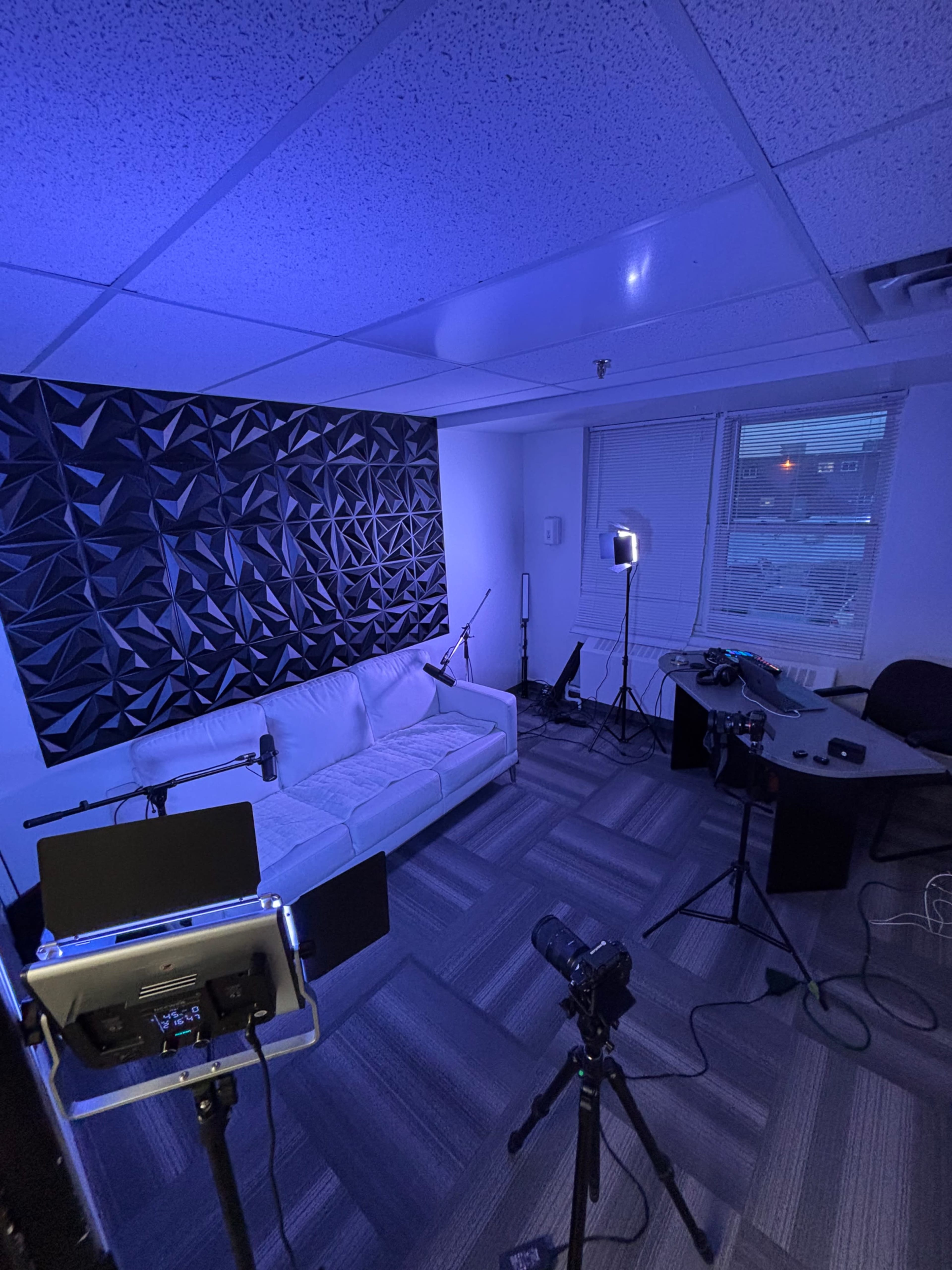 The image shows a small room equipped with a white couch, a geometric patterned wall, and various cameras and lighting equipment set up for a video production.