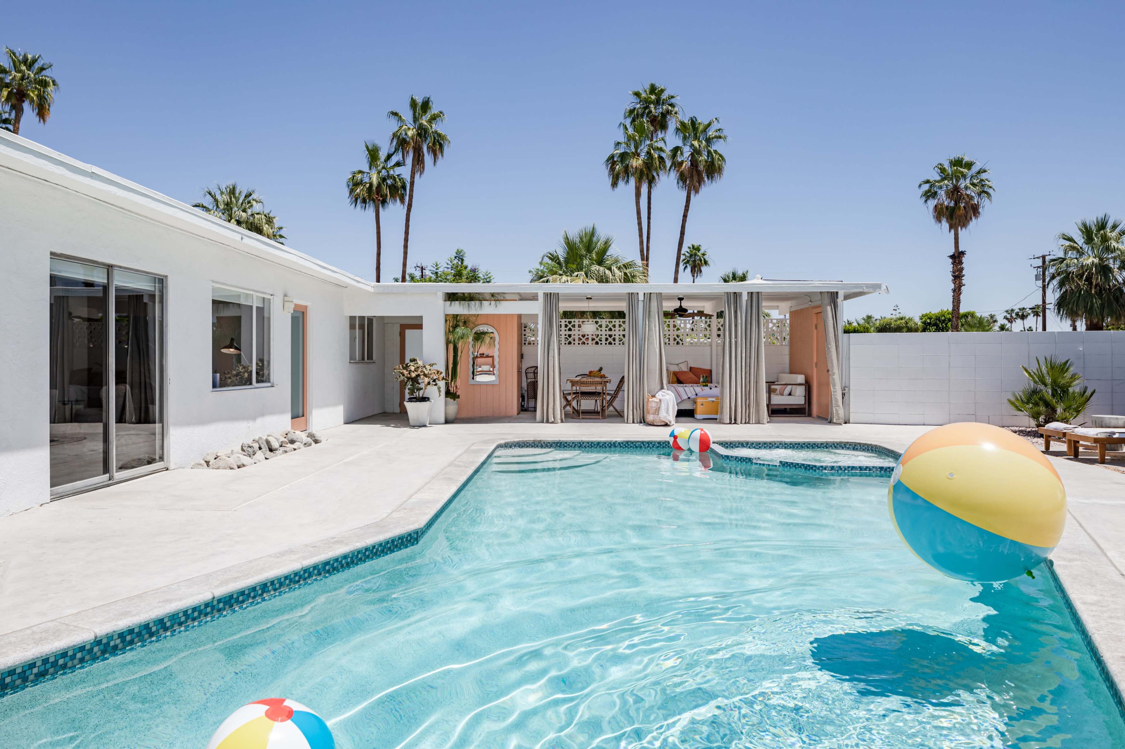 A swimming pool with colorful beach balls is surrounded by a modern house and palm trees under a clear sky.