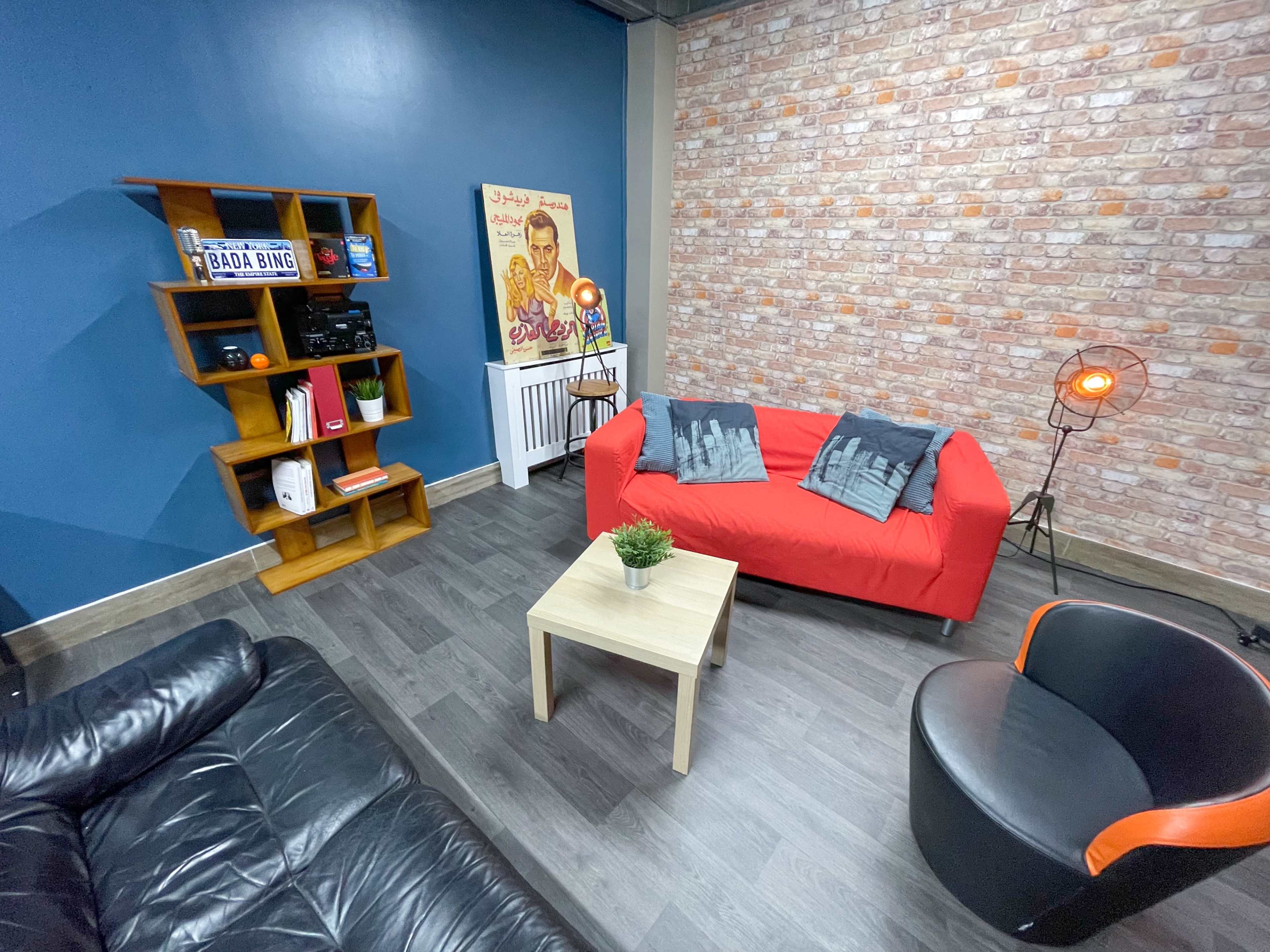 A cozy room with a red sofa, a black leather couch, a small wooden table, and a decorative wall featuring a brick design and a vintage poster.