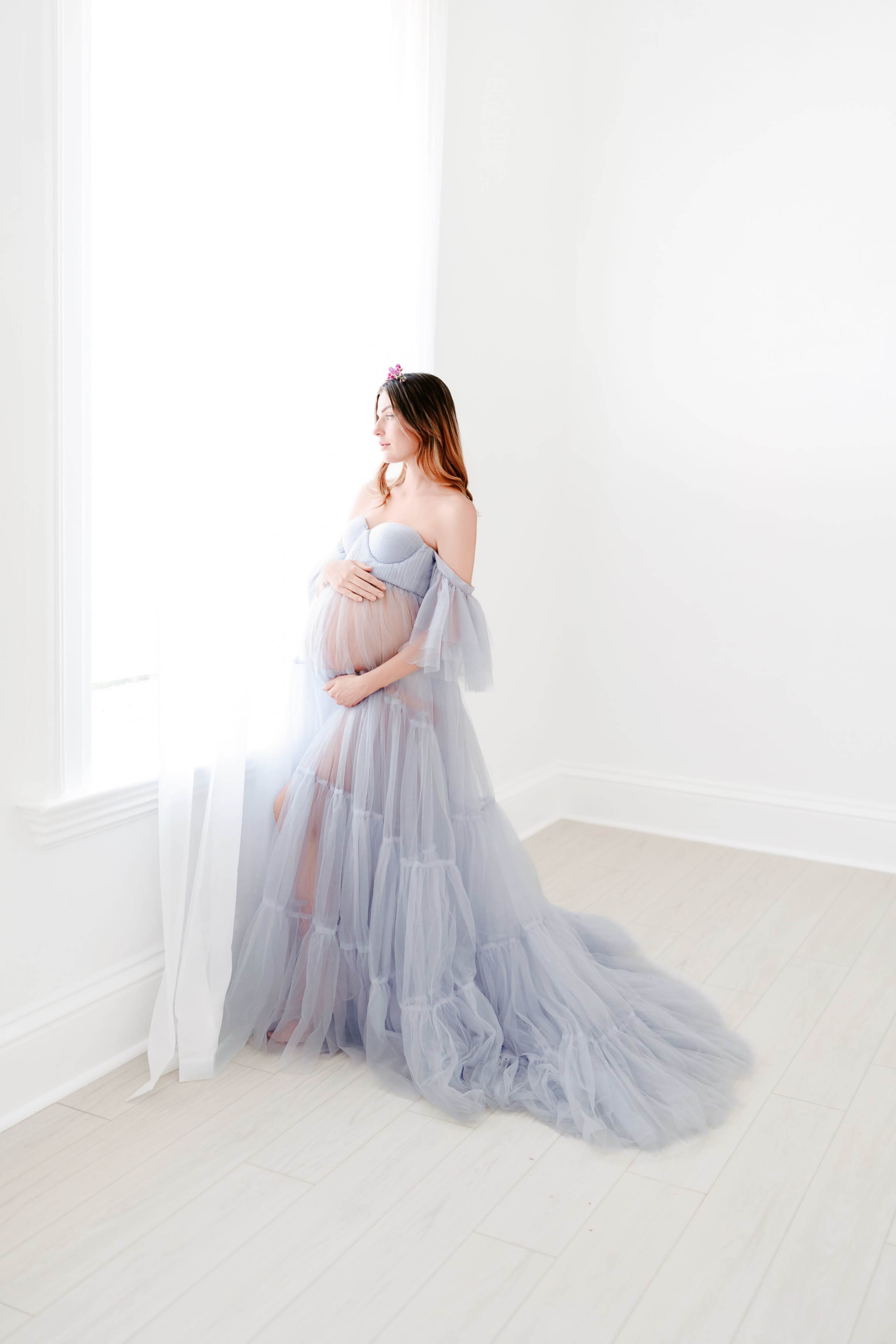 A pregnant woman in a flowing, gray tulle gown stands by a window, gently cradling her belly.