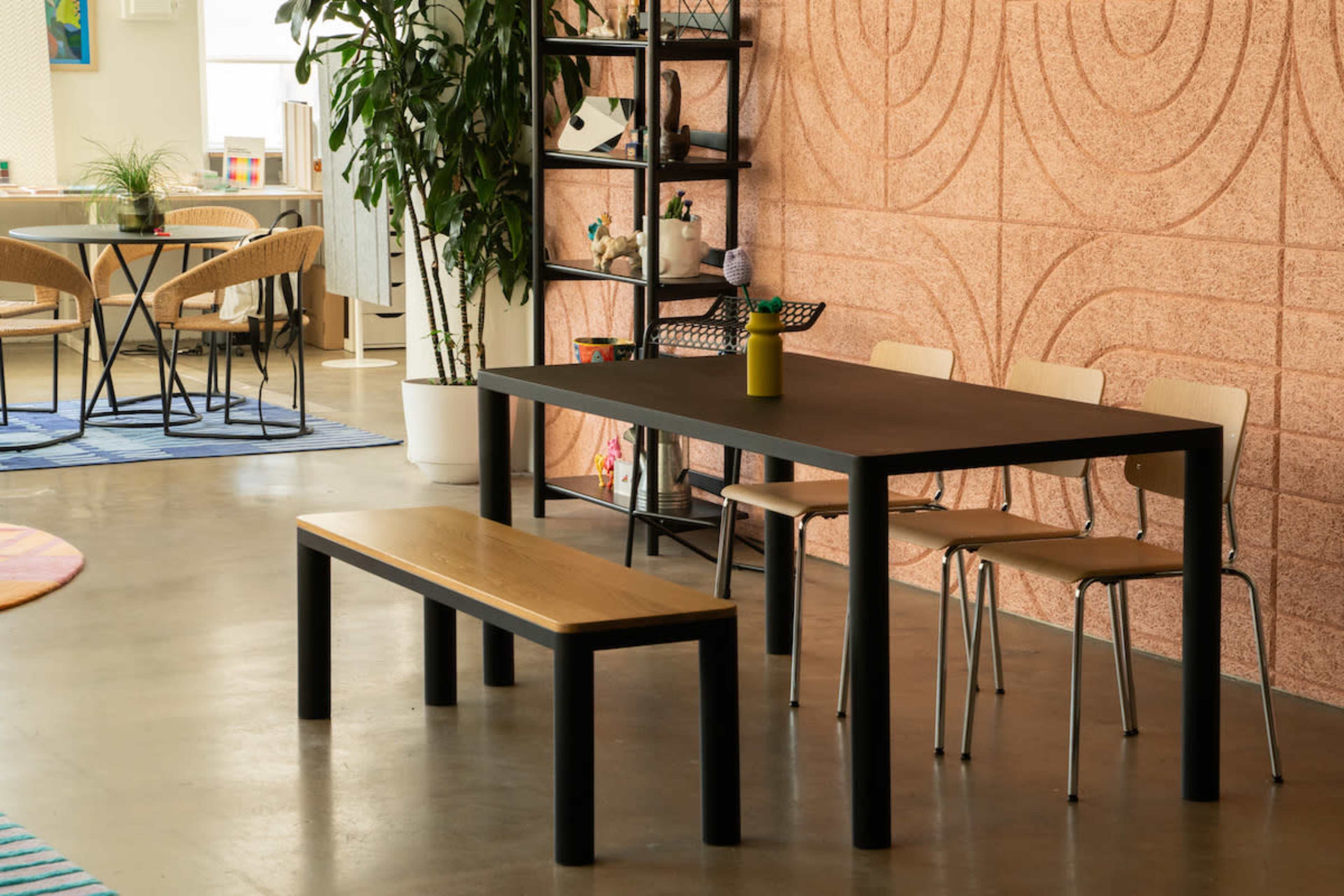 A black table with a wooden bench and chairs is set in a modern interior featuring textured walls and plants.