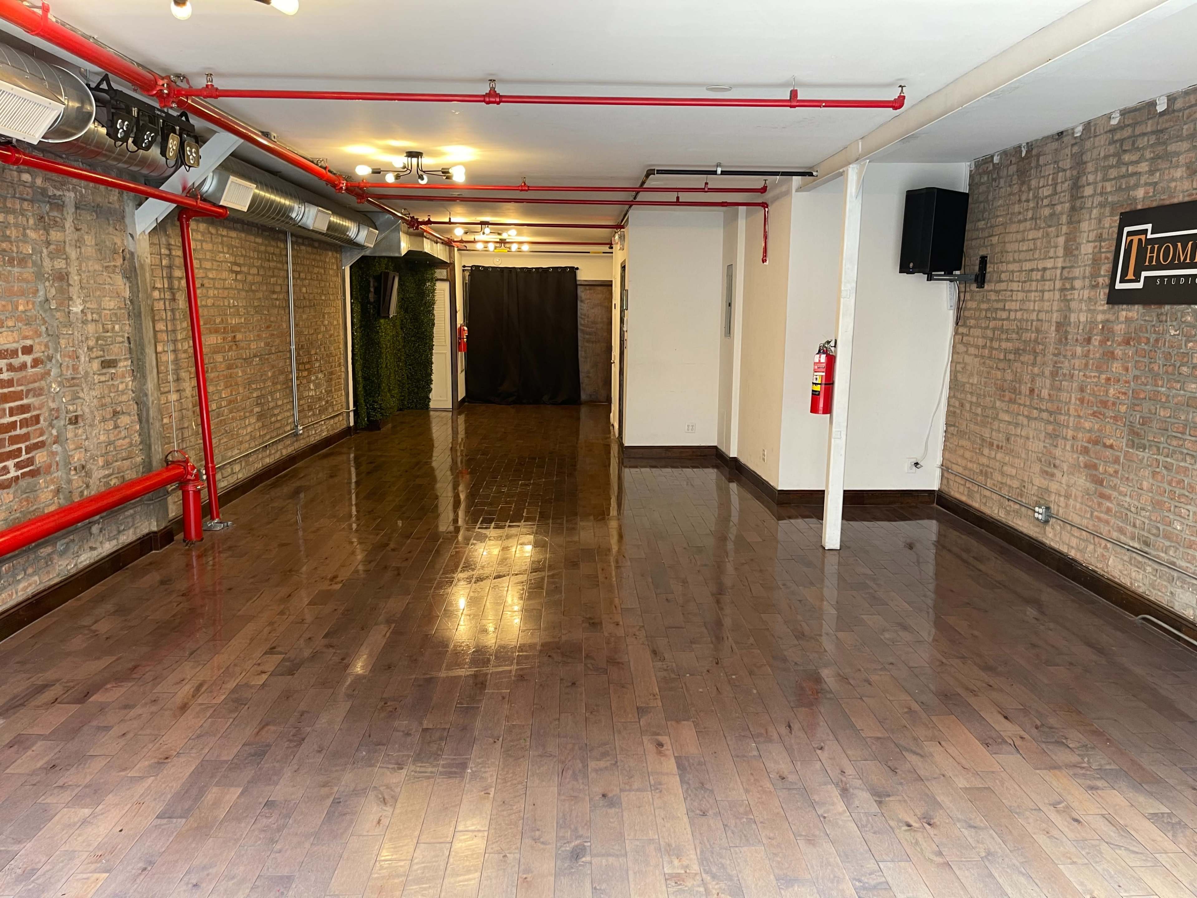An empty room with polished wooden floors, exposed brick walls, and red pipes along the ceiling.
