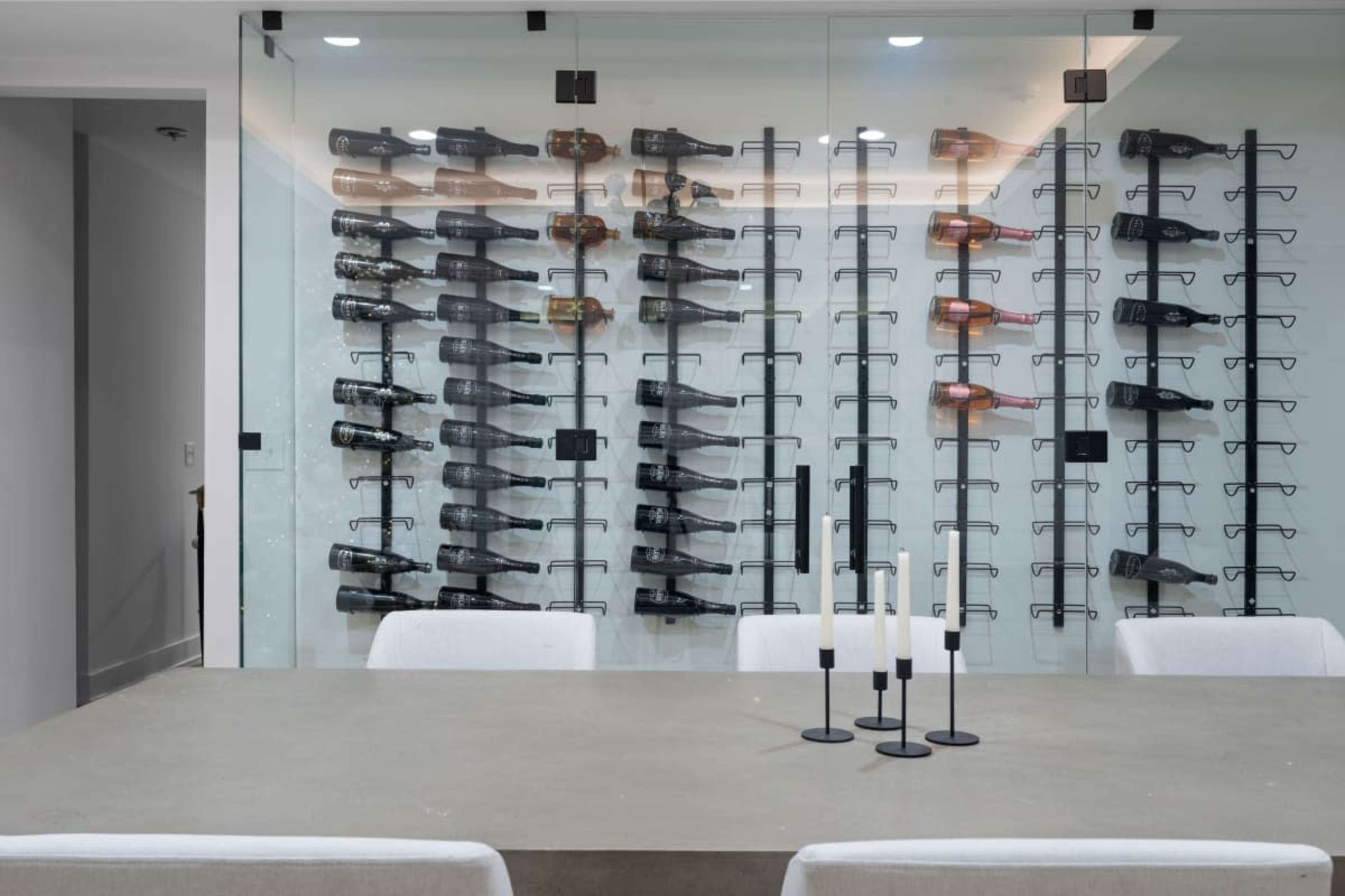 The image shows a modern dining area with a concrete table, white chairs, and a glass wall displaying numerous wine bottles arranged on black racks.
