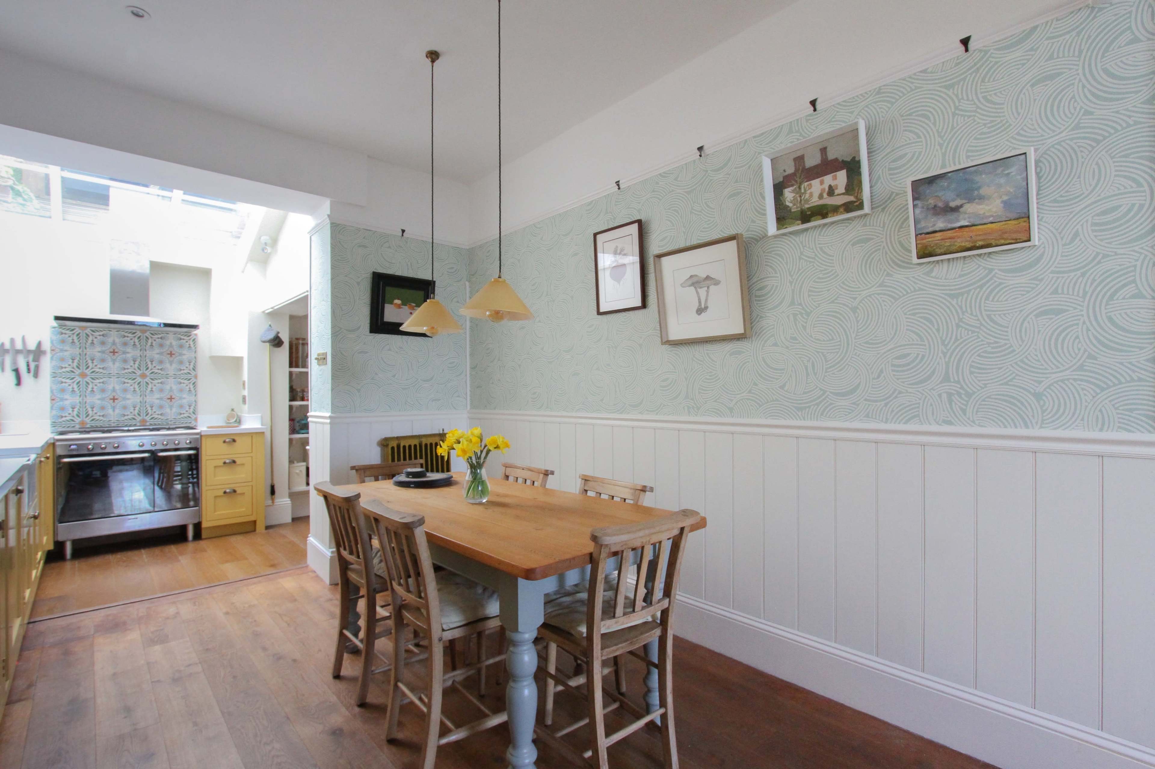 A dining area features a wooden table with chairs, yellow flowers in a vase, and patterned wallpaper, leading into a kitchen space with a stove and colorful tile backsplash.