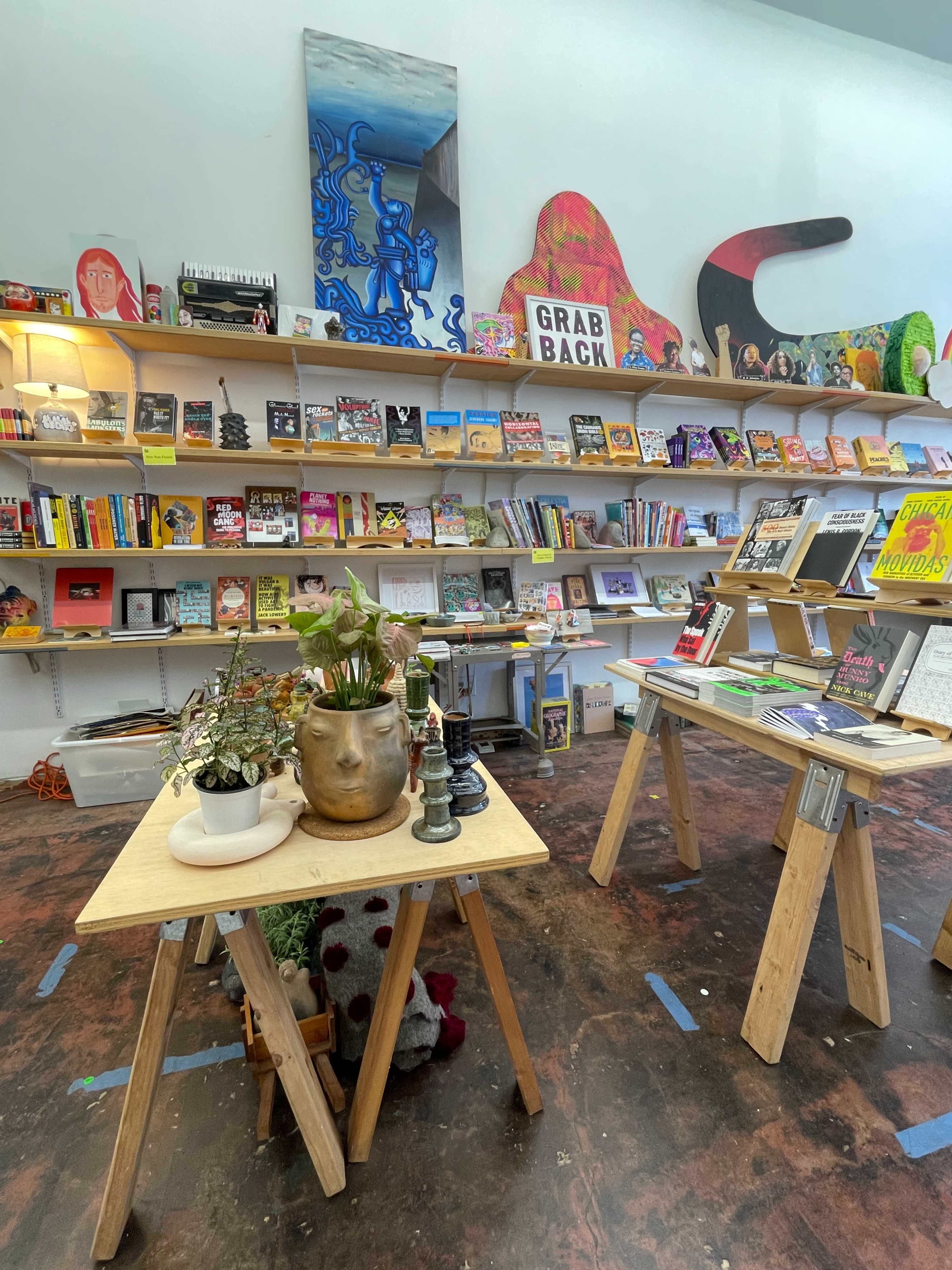 The image shows a colorful bookstore interior with wooden tables displaying plants and books, along with shelves filled with various titles and art pieces on the walls.