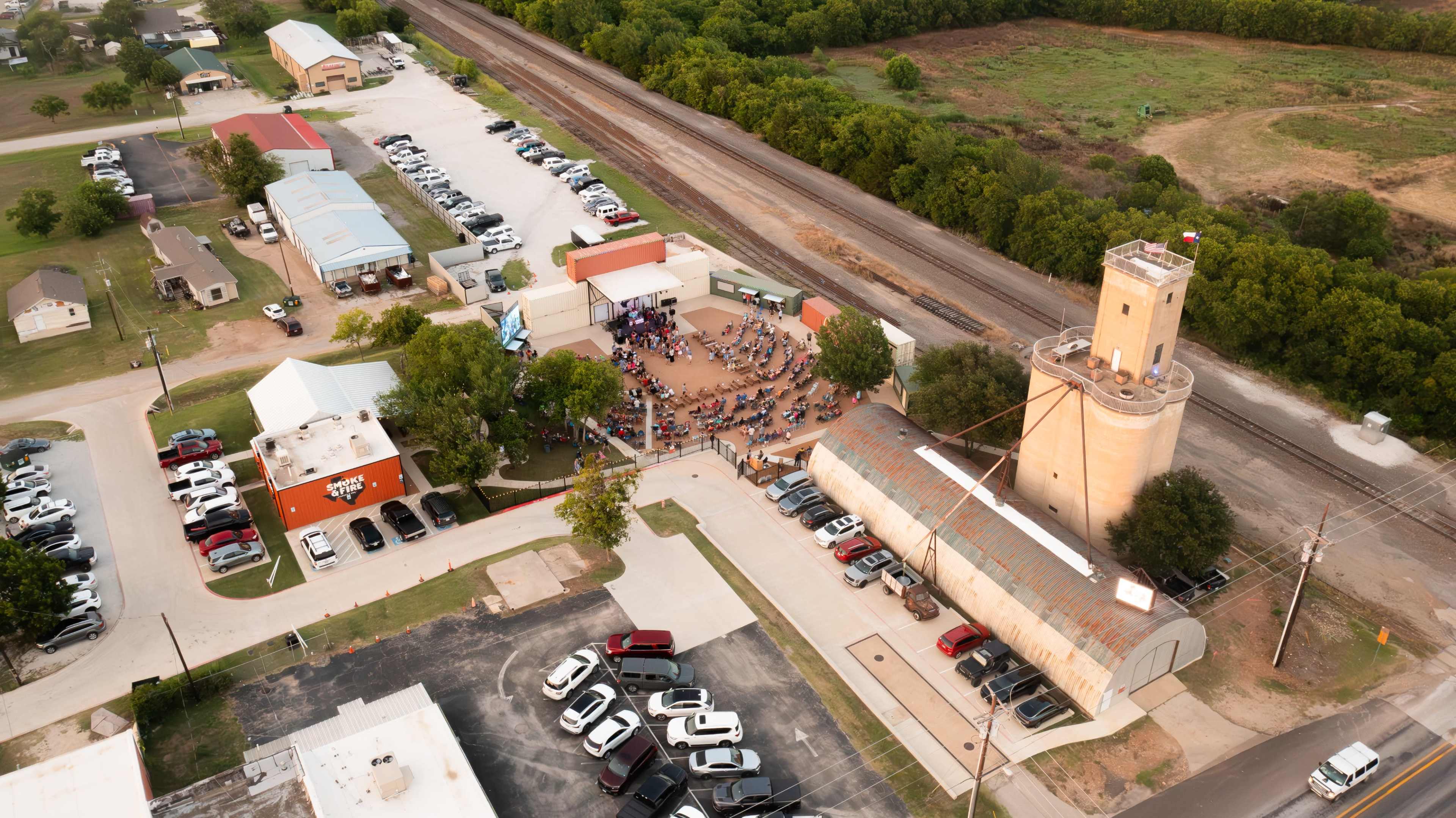 Aerial view of a gathering area with multiple buildings and parked cars near a railroad track and green fields.