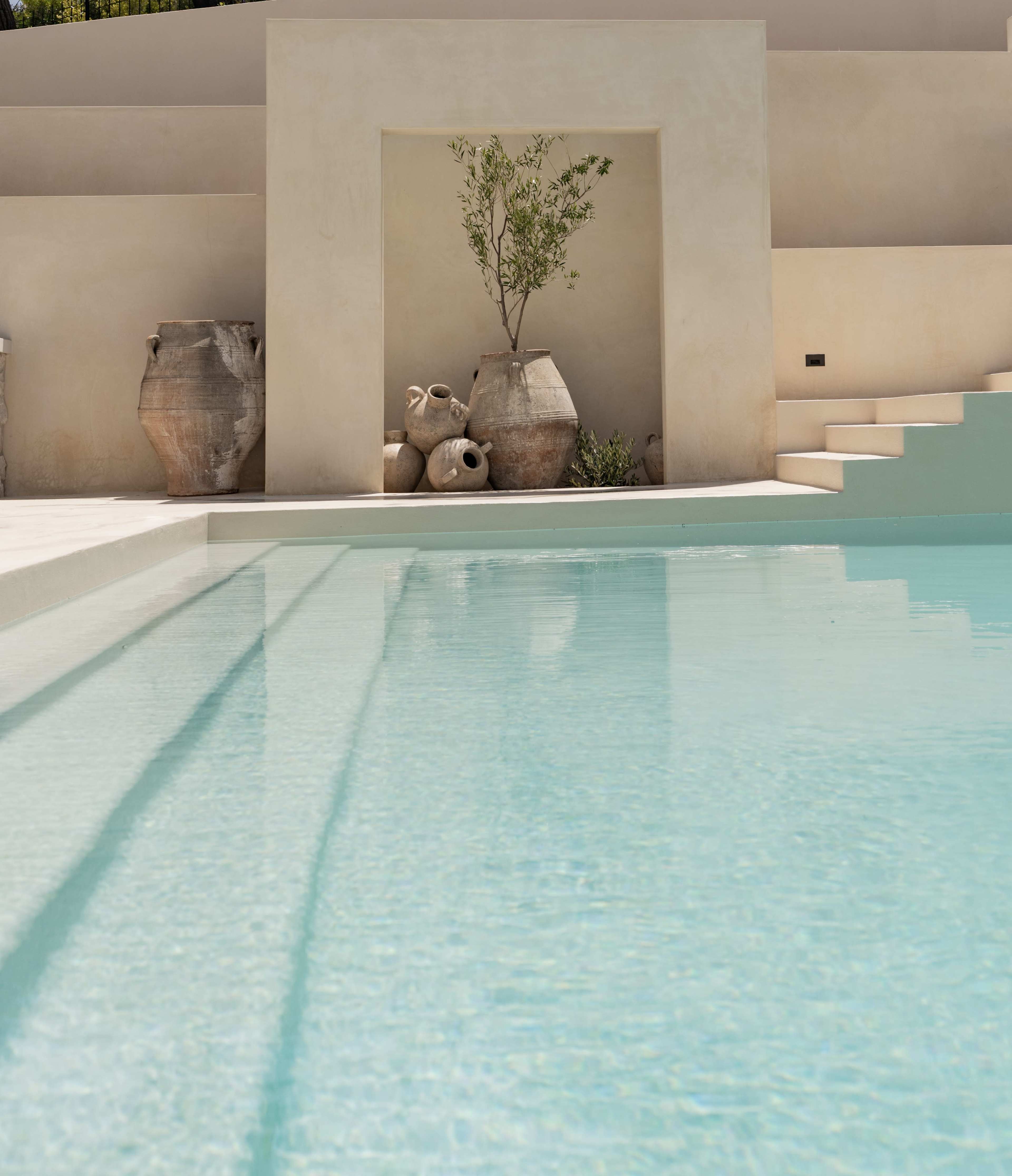 The image shows a clear swimming pool with shallow steps leading to a textured wall adorned with large clay pots and a small plant.