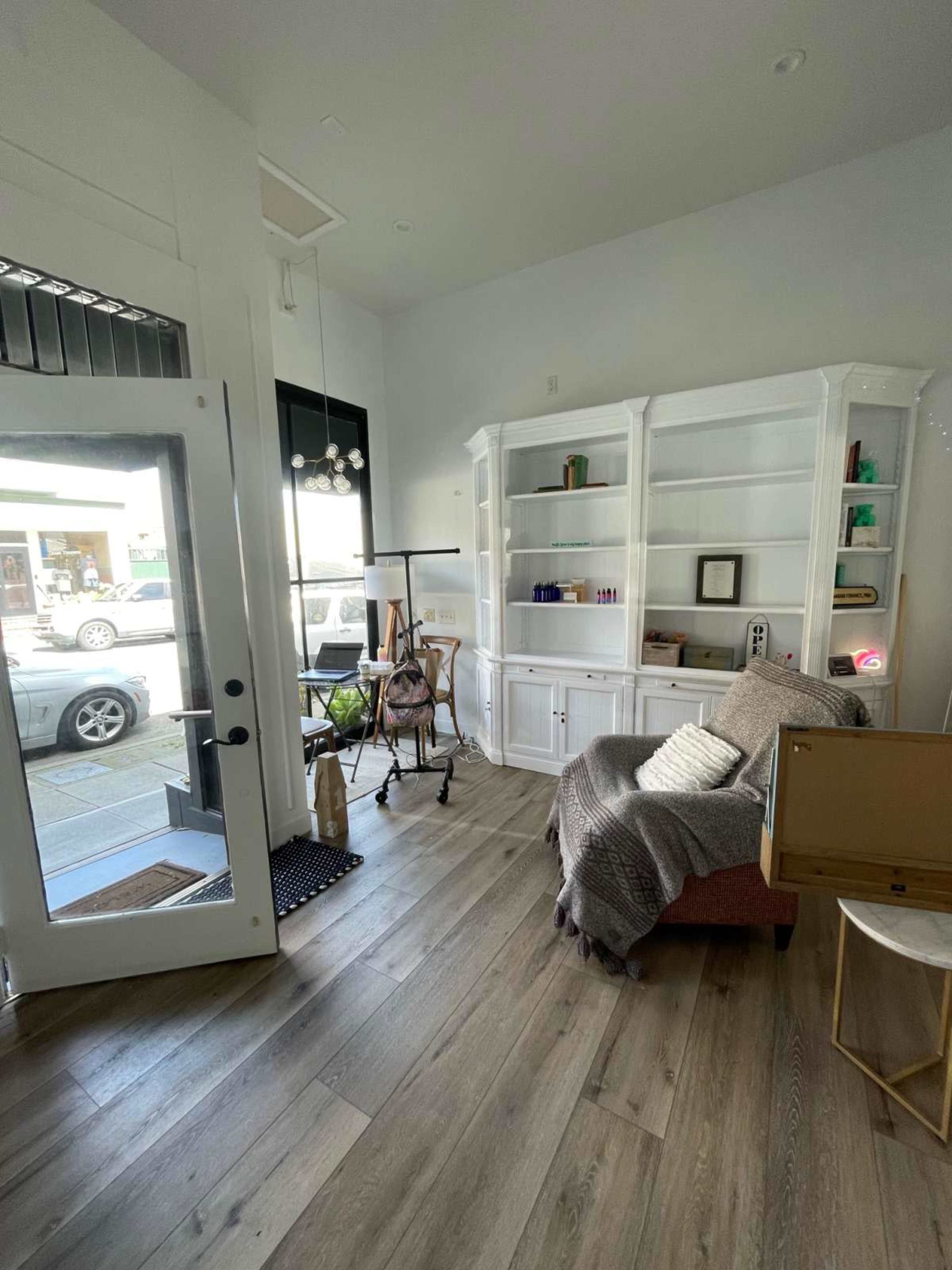 A bright interior of a room featuring a white bookshelf, a cozy chair draped with a blanket, and a large front window with a view of parked cars outside.
