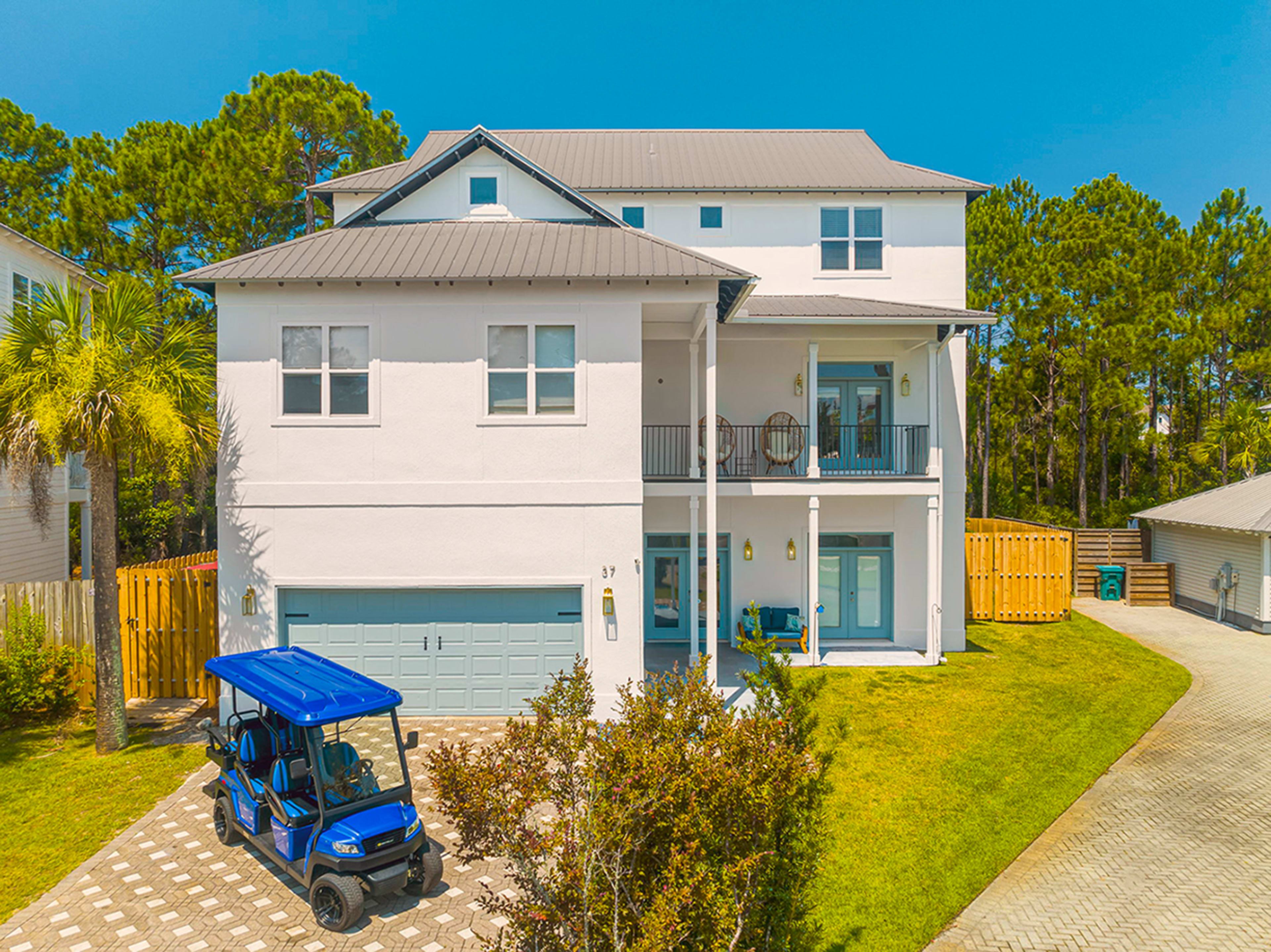 A two-story, modern white house with a gray metal roof, a paved driveway, a parked blue golf cart, and surrounded by trees.