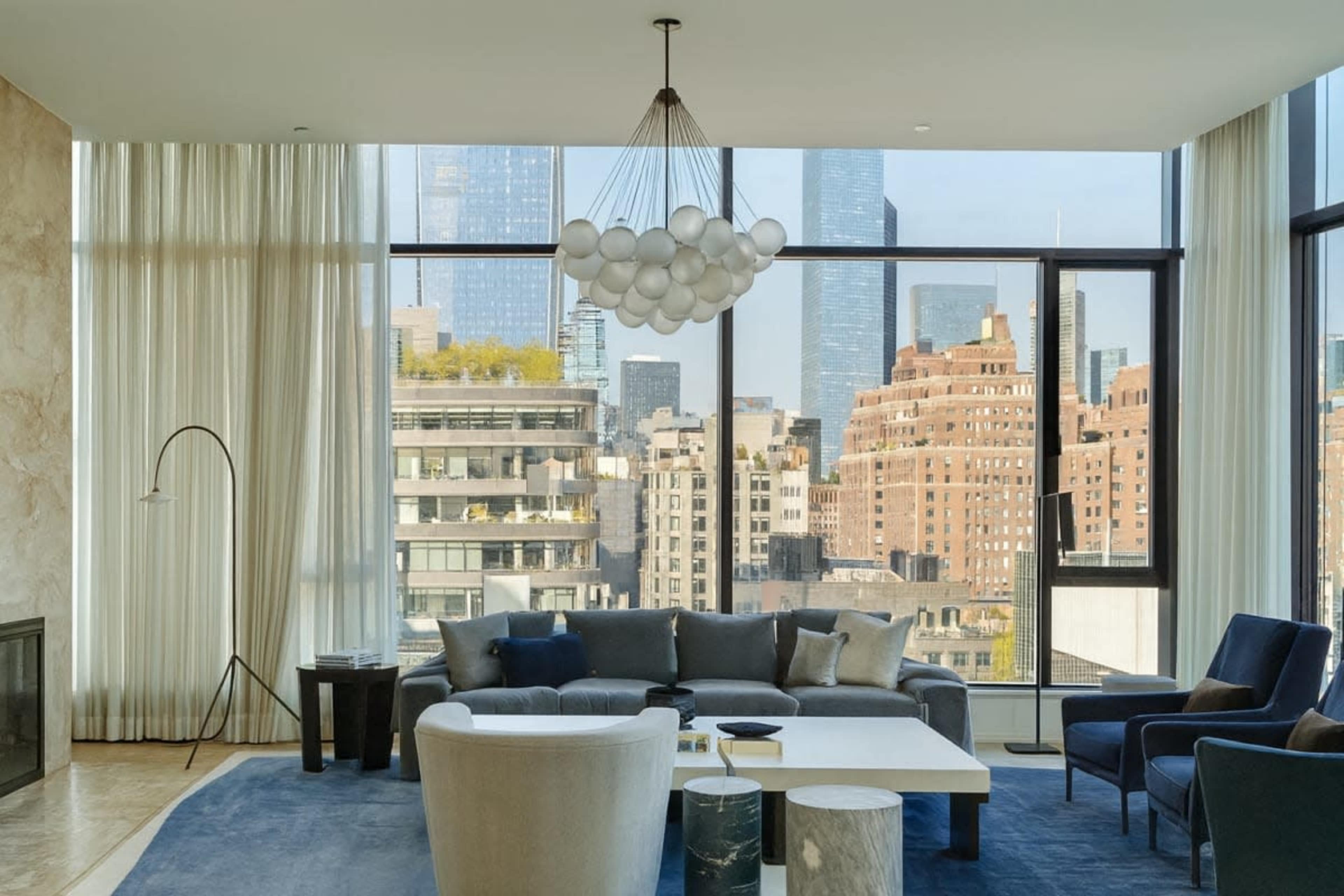 The image shows a modern living room with large windows revealing a city skyline, featuring a seating area with a sectional sofa, armchairs, and a decorative light fixture.