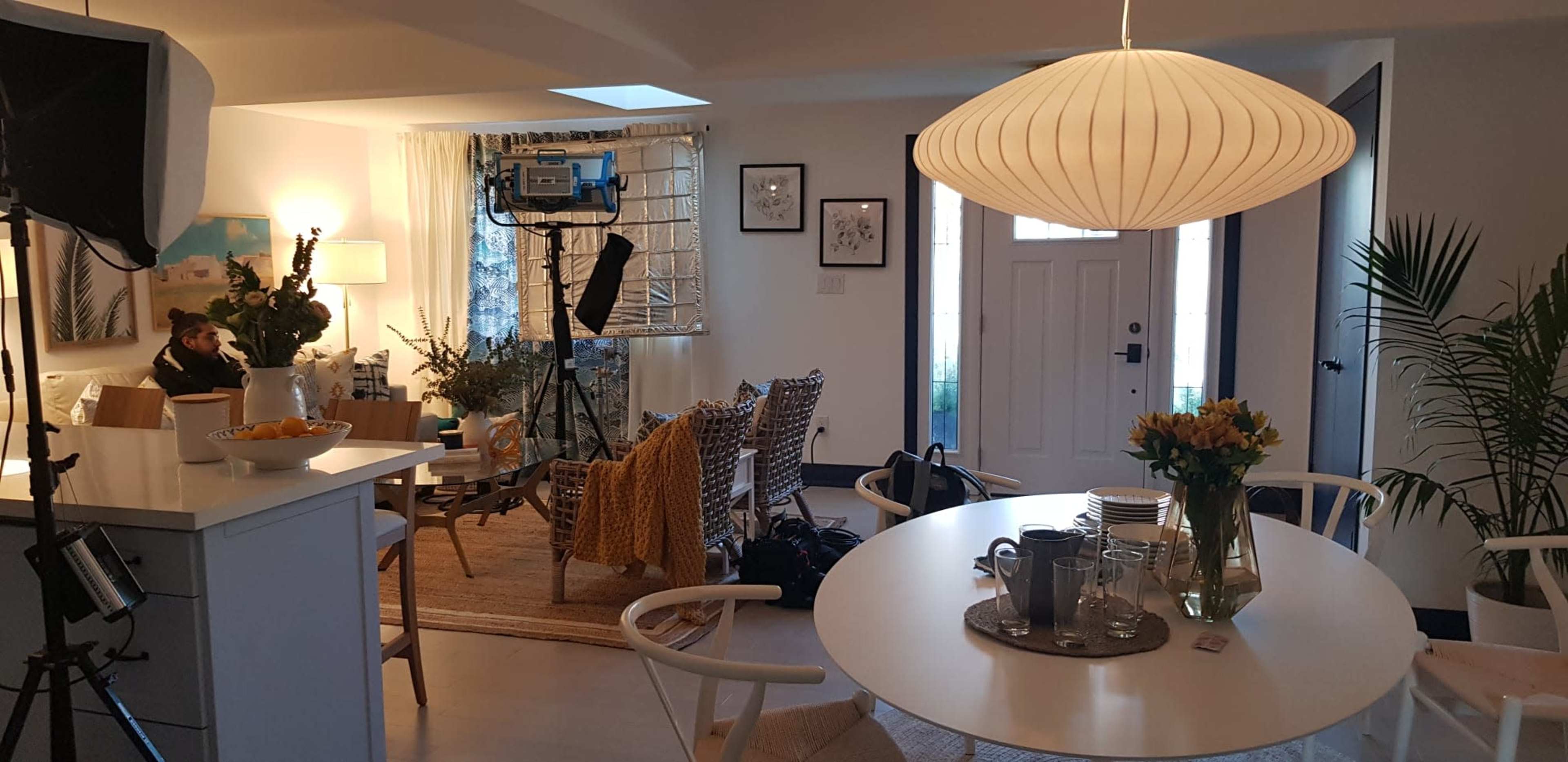 The image shows a brightly lit indoor living space set up for a filming shoot, featuring a dining table with tableware, a cozy seating area with decorative elements, and a light fixture overhead.