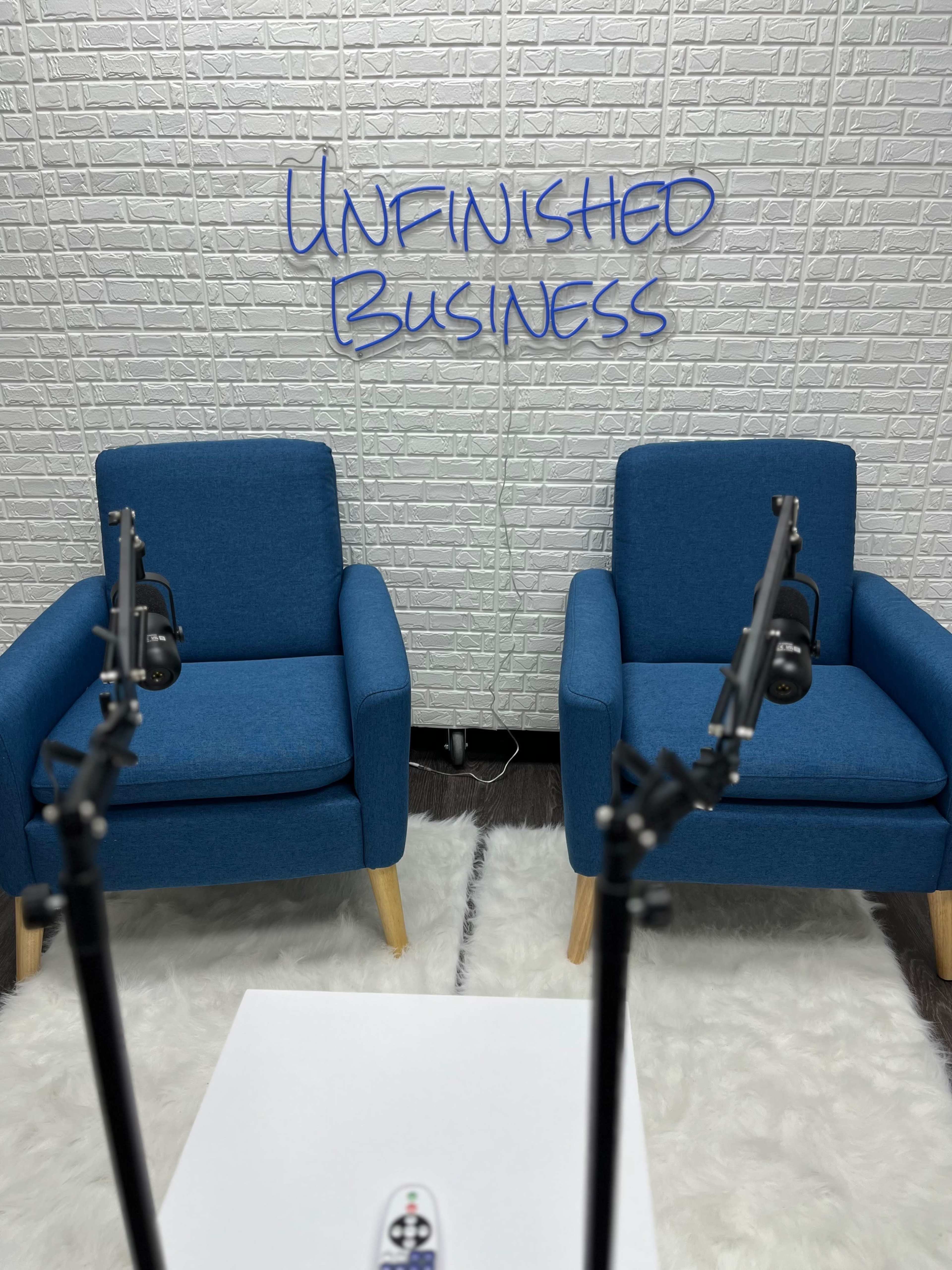 A white brick wall features the phrase "UNFINISHED BUSINESS" above two blue chairs positioned across from each other, with microphone stands in the foreground.