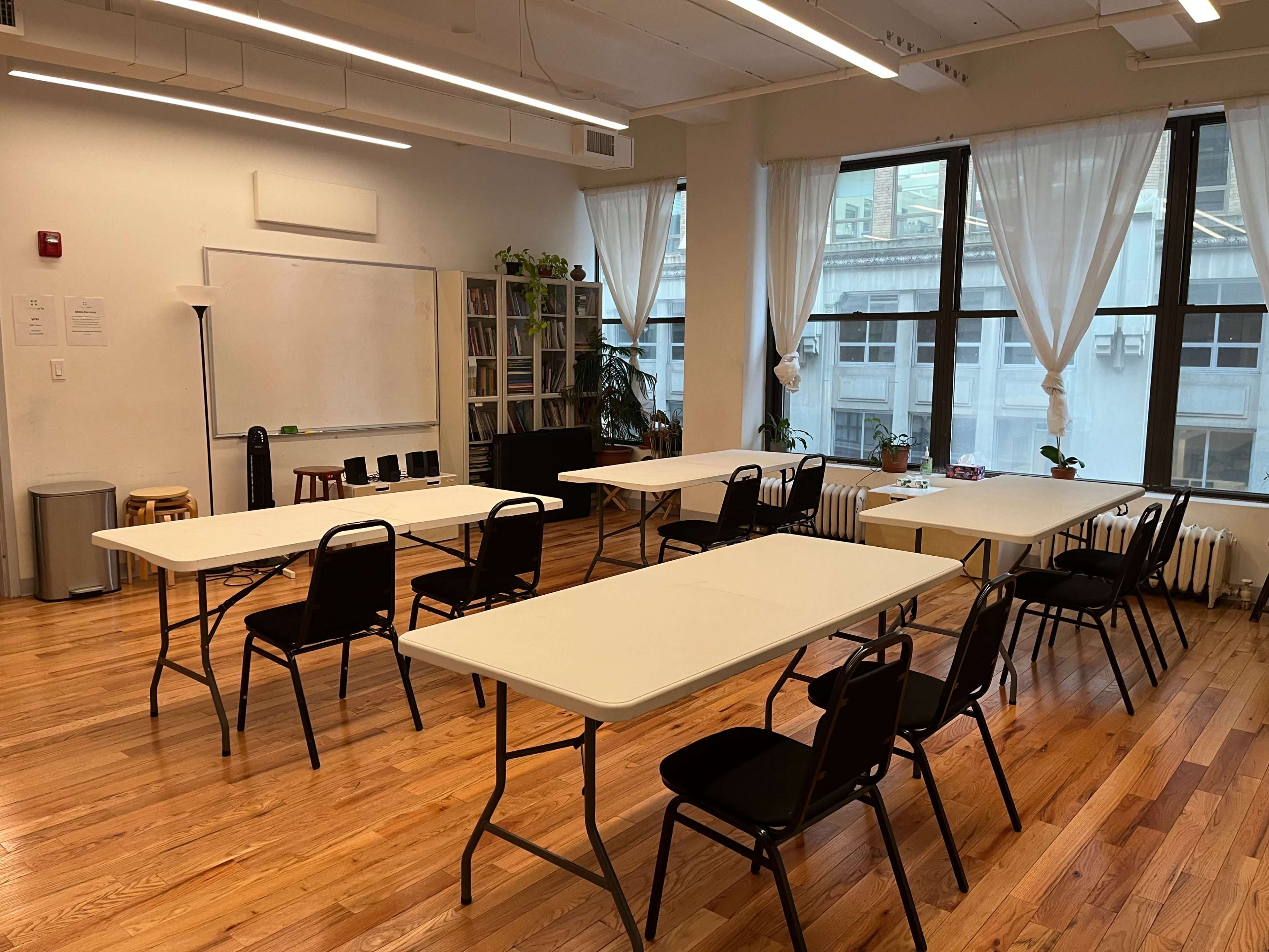A bright, spacious classroom features several folding tables arranged in a diagonal pattern, with chairs placed around them and large windows allowing natural light.