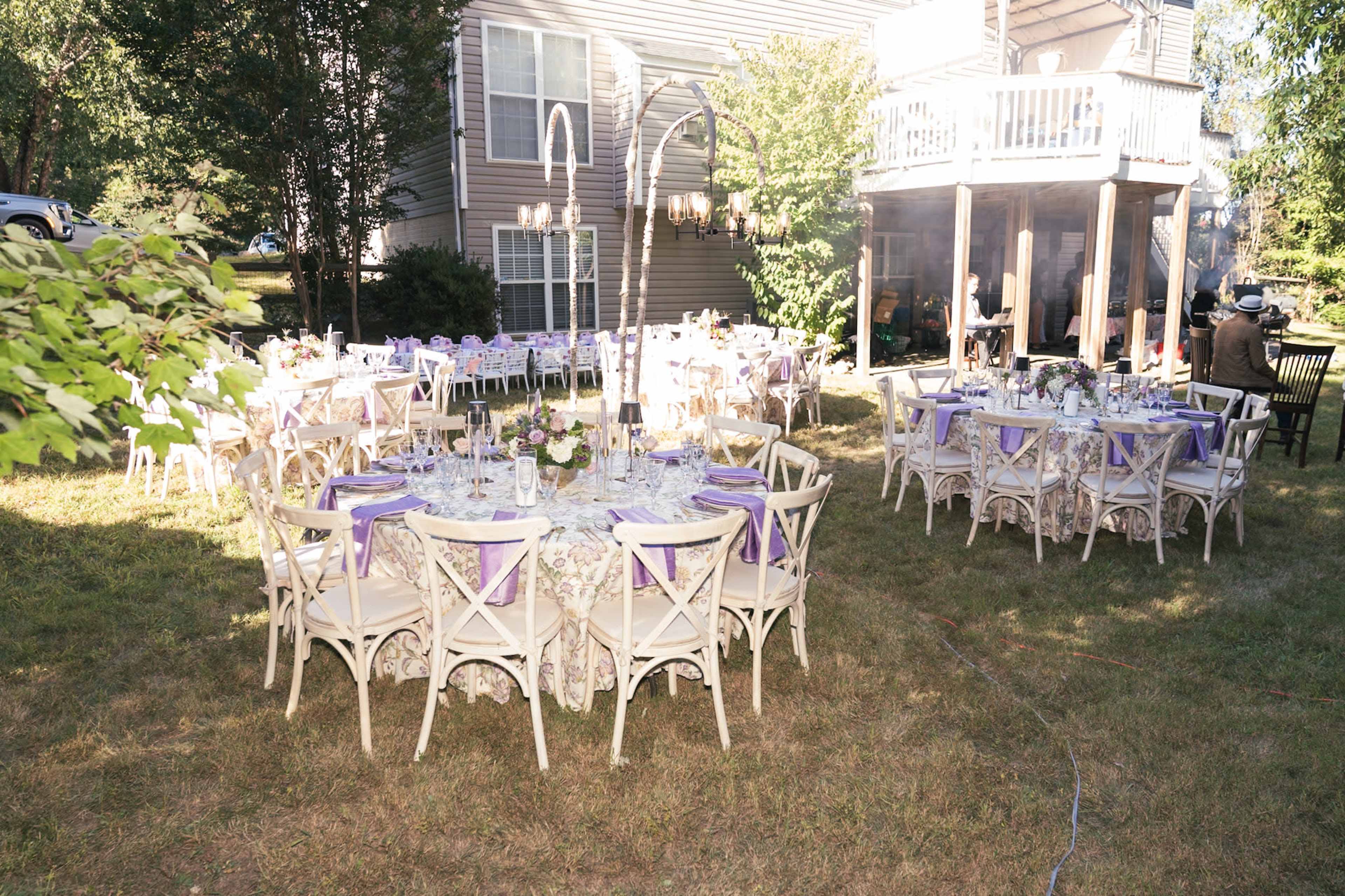The image shows a backyard setting with several round tables covered with purple accents, arranged for a formal outdoor event near a house.