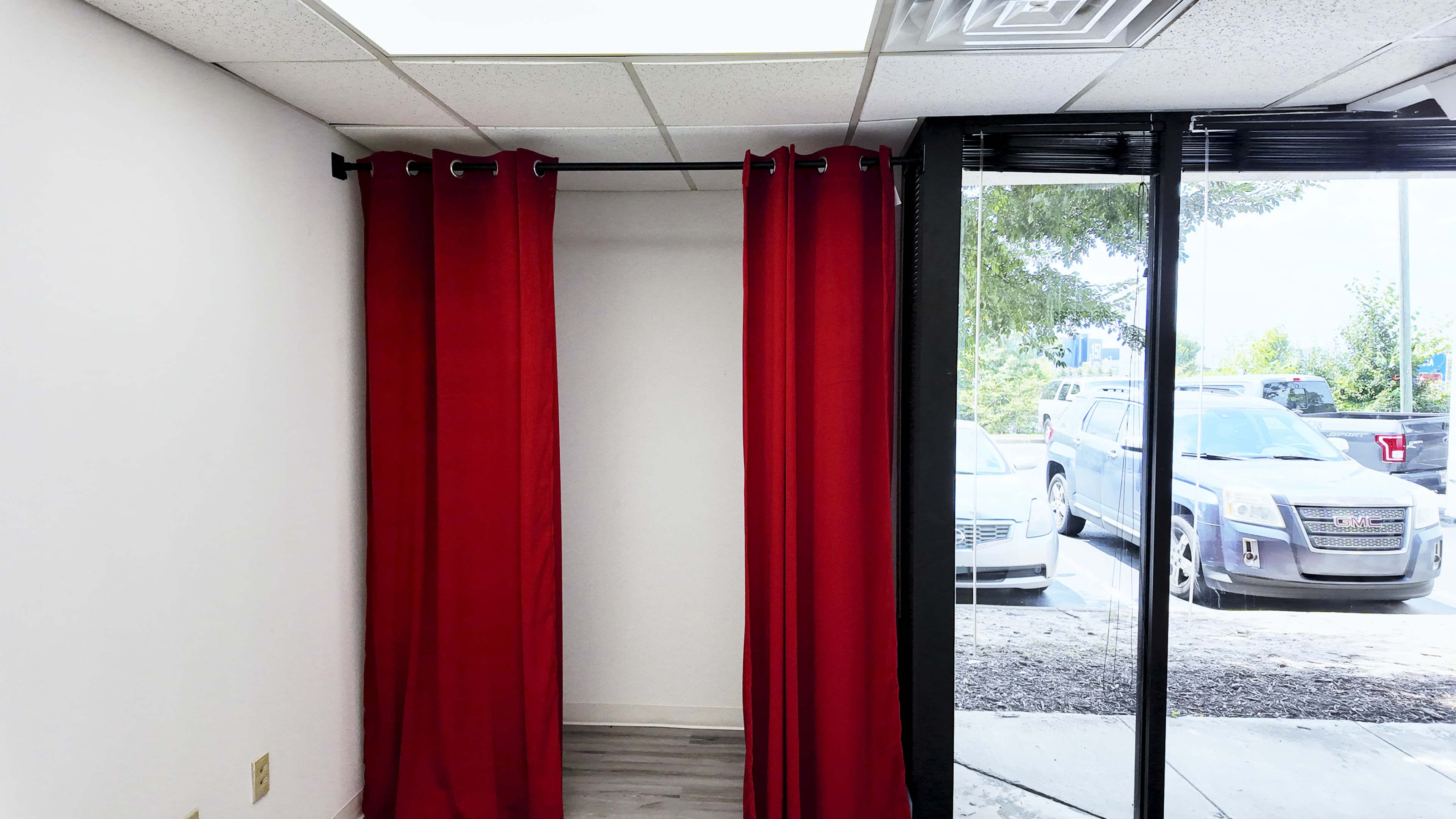 An empty room with red curtains hanging on a rod, and large windows revealing parked cars outside.