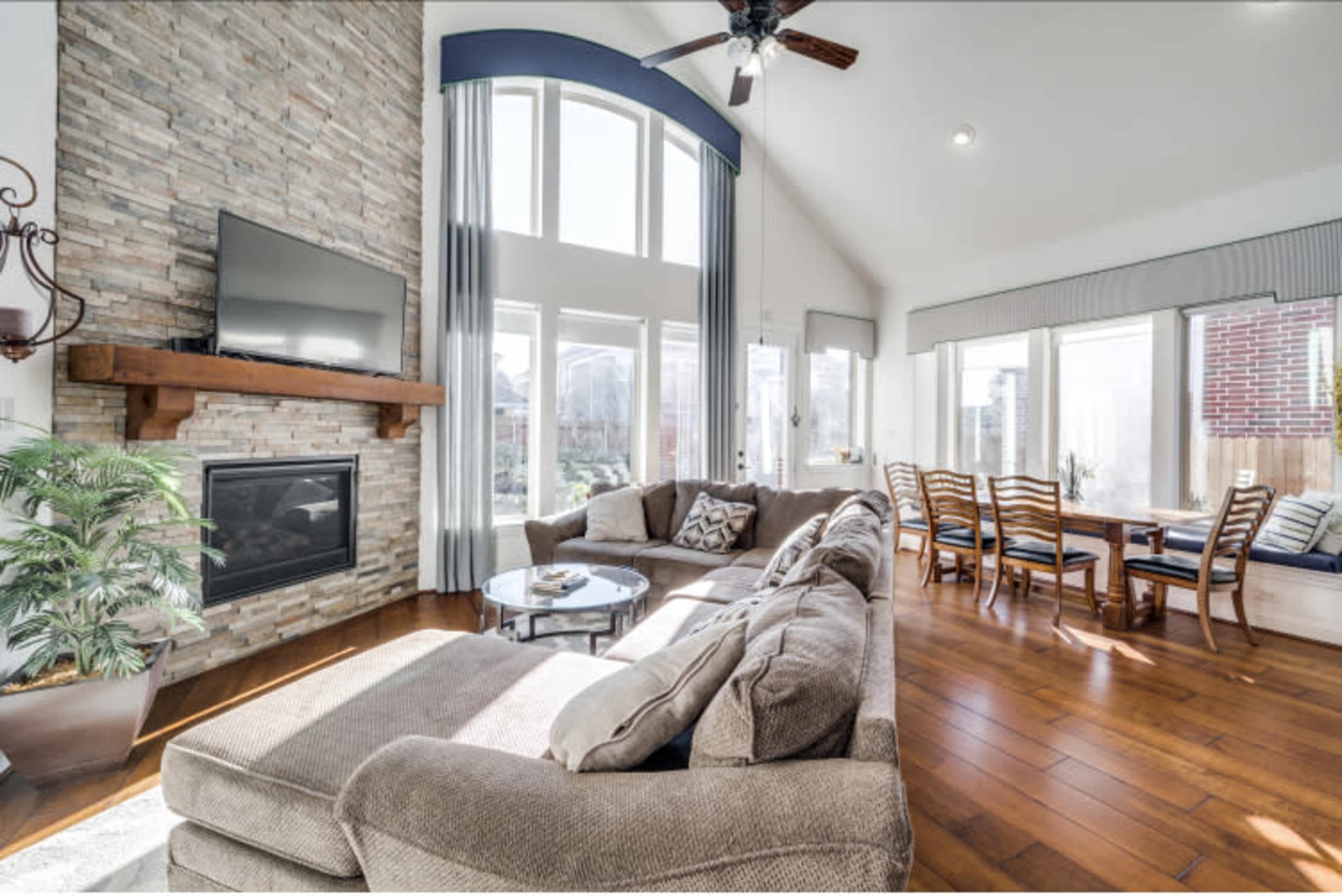 A spacious living room features a stone fireplace with a mounted TV, large windows allowing natural light, and a comfortable sectional sofa facing a dining nook.