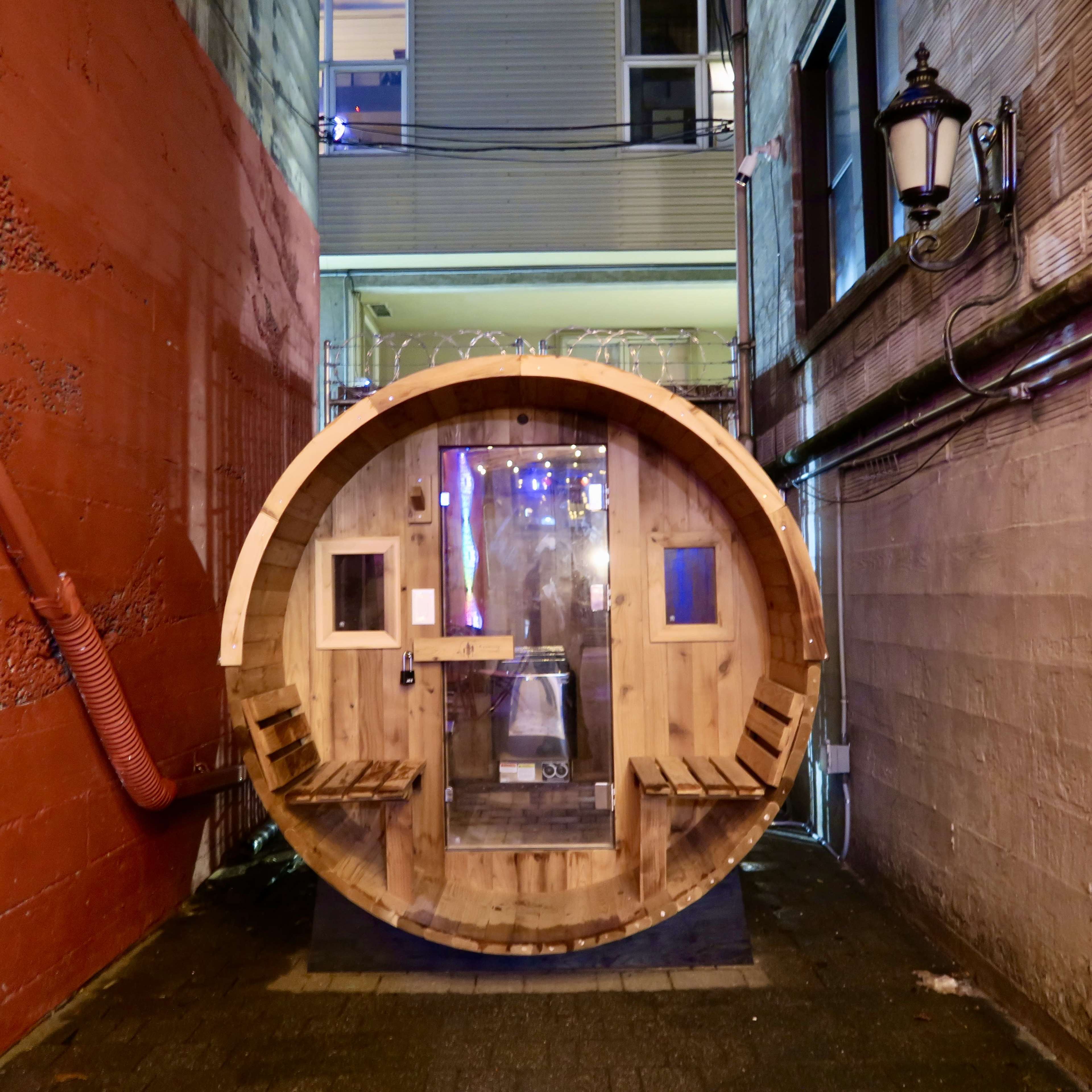 A circular wooden structure with two benches stands in an alleyway, illuminated by ambient light from a nearby building.