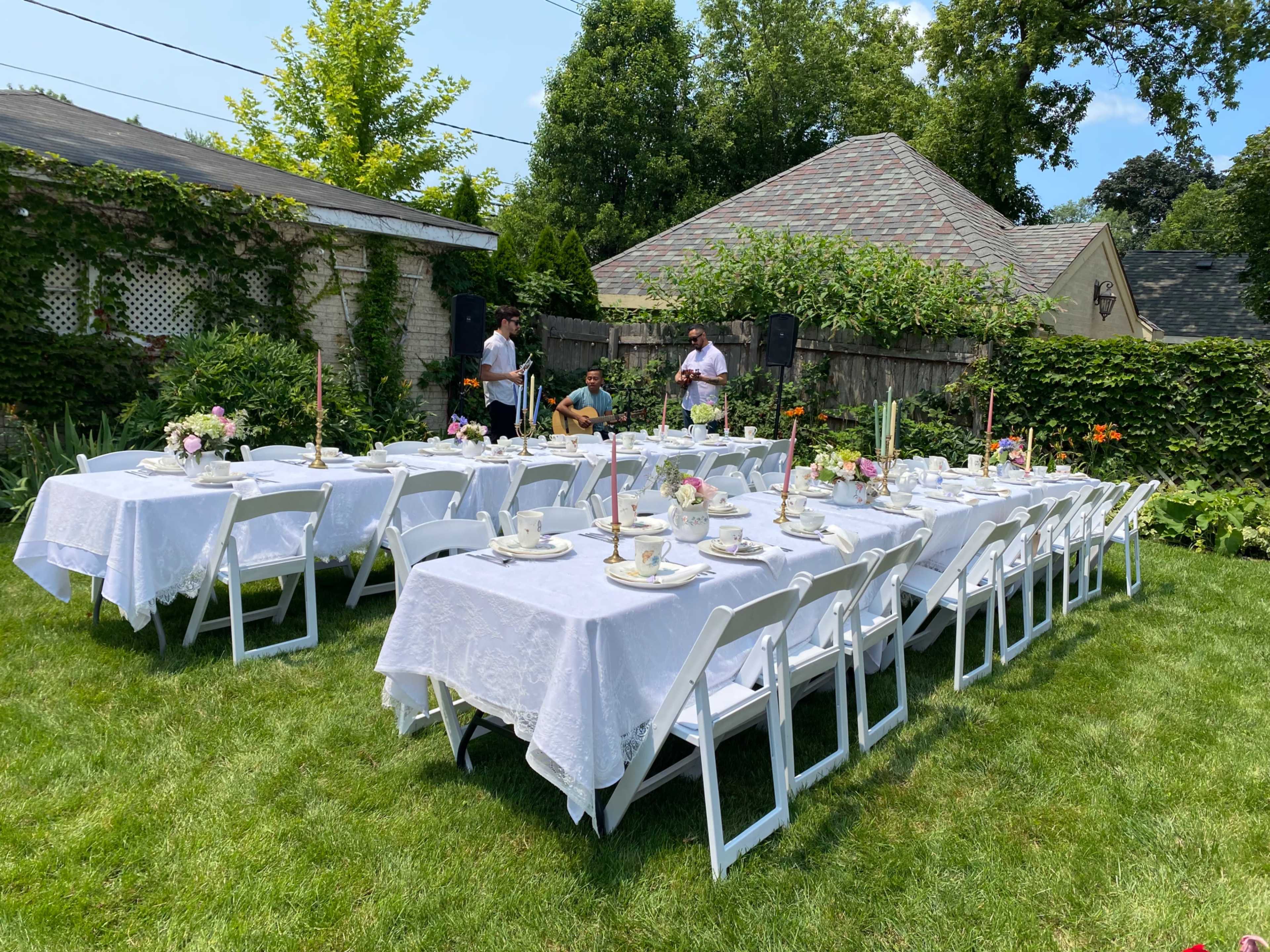 A garden is set up with multiple white tables covered in lace tablecloths, each adorned with floral centerpieces and arranged for a gathering.