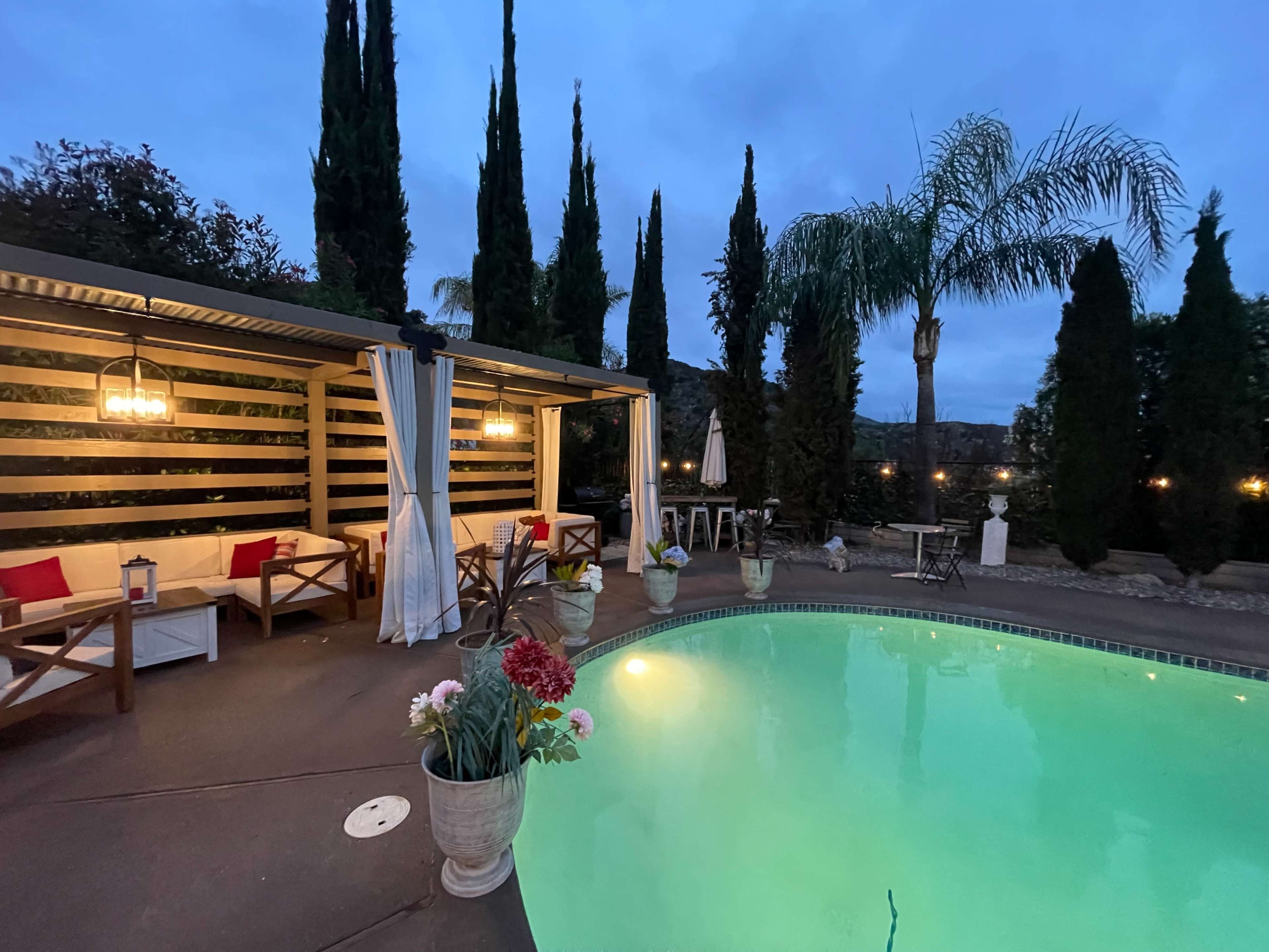 A swimming pool surrounded by tall greenery and a shaded seating area with lanterns, set against a twilight sky.