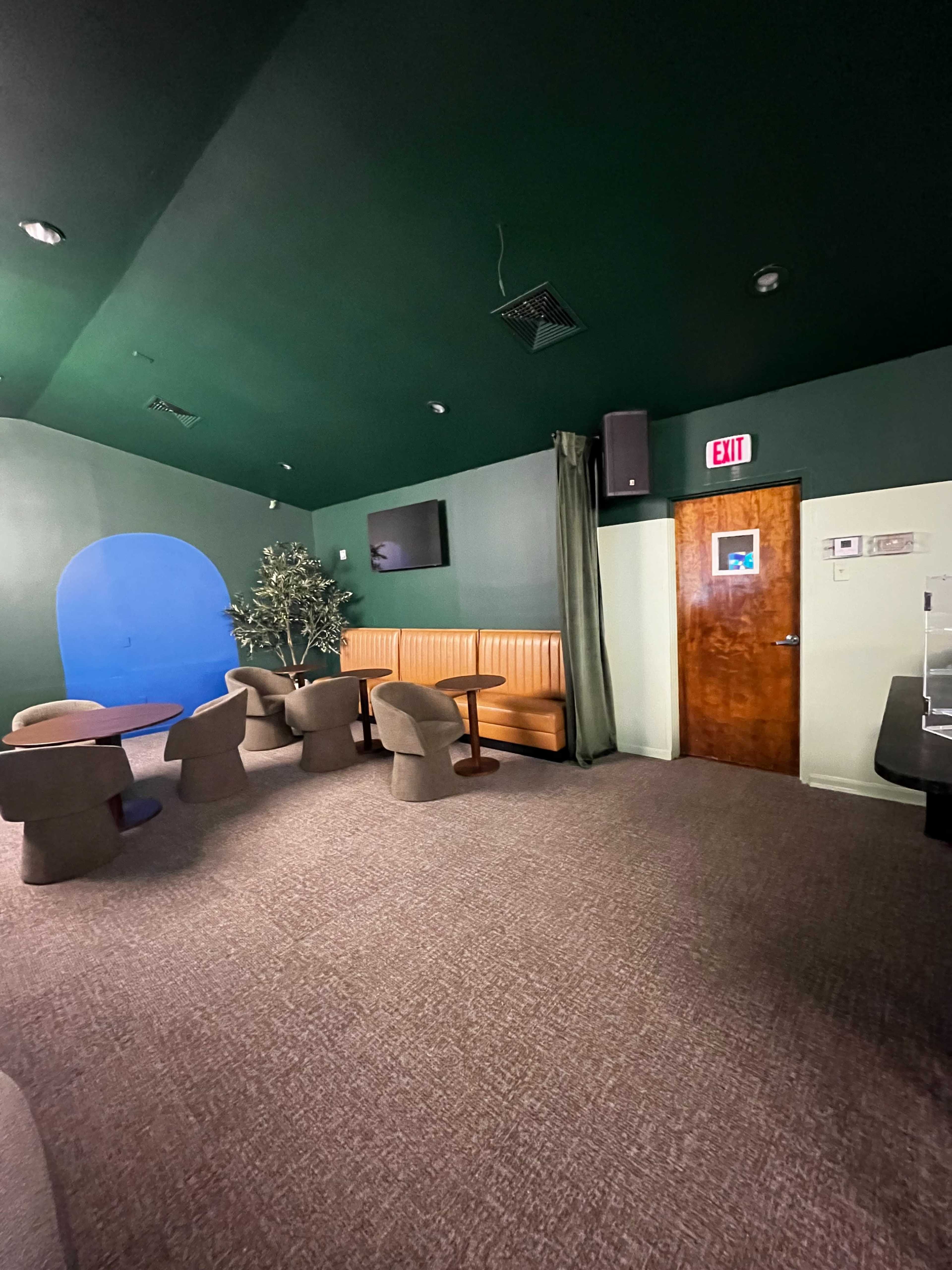The image shows a lounge area with orange booth seating, several brown chairs, a wooden door, and a dark green ceiling.