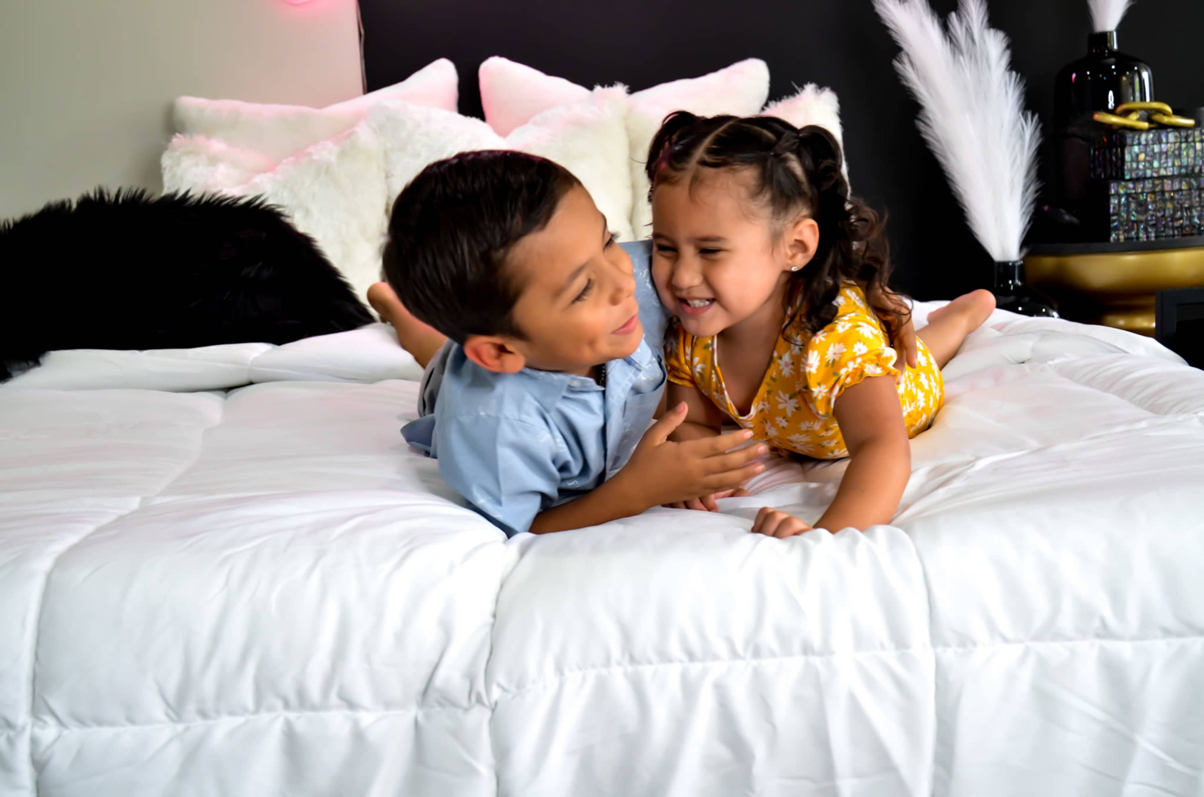 Two children lie on a white bed, smiling and playfully leaning towards each other.