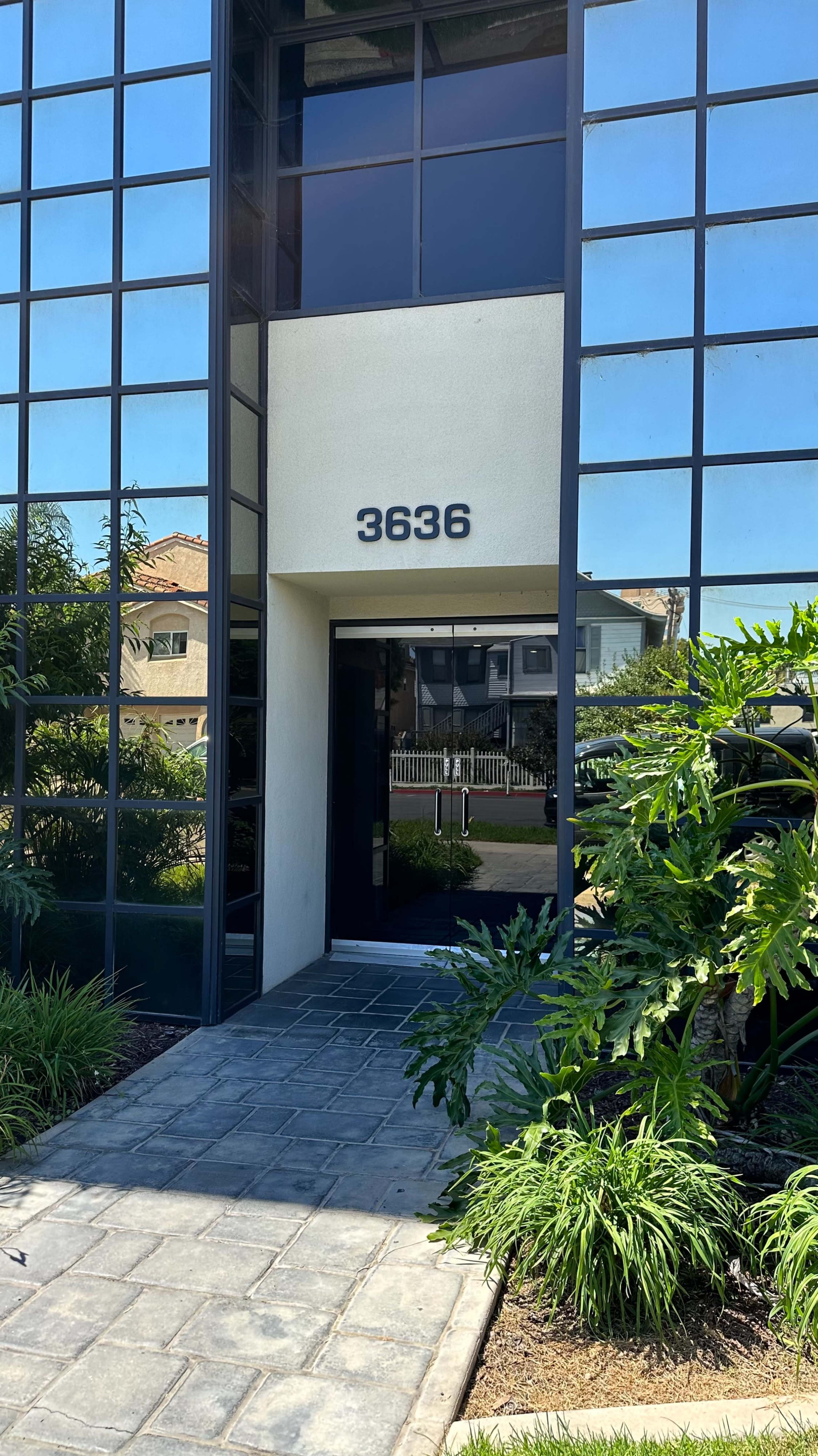 A modern building entrance with large glass windows, featuring the number "3636" prominently displayed above the door and surrounded by landscaped greenery.