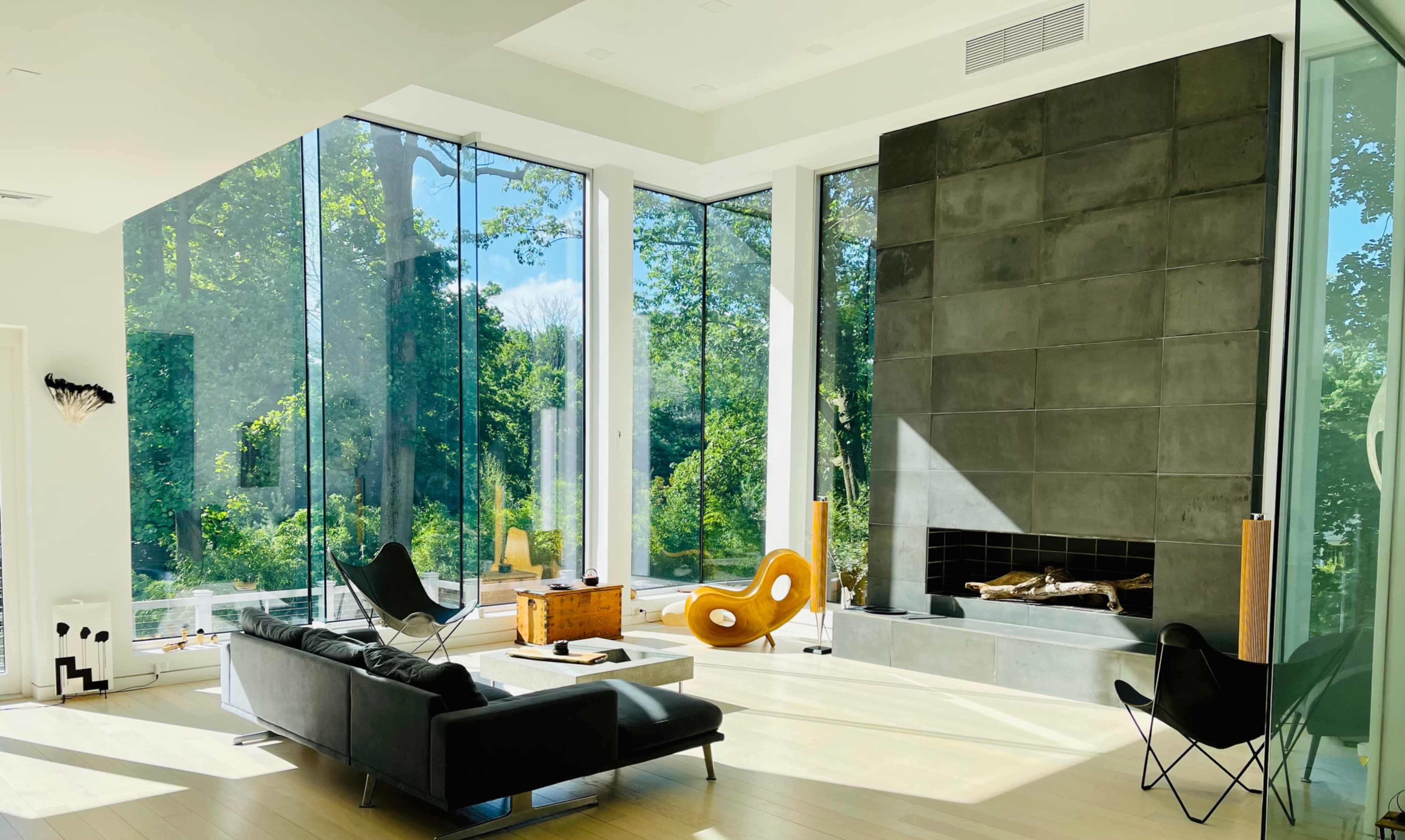 The interior of a modern living room features large windows overlooking a garden, a stone fireplace, and minimalist furniture including a black couch and a unique orange chair.