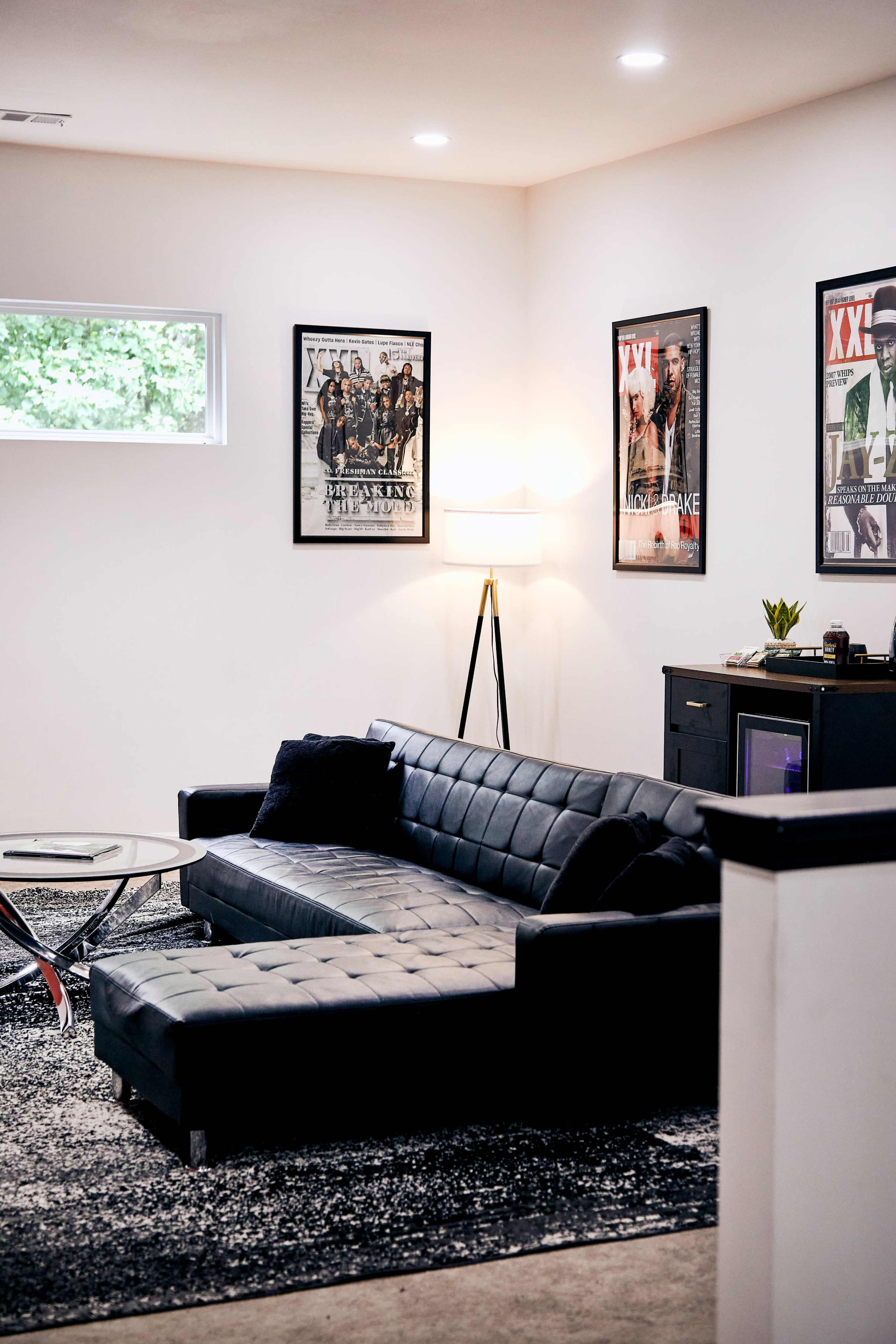 A modern living room features a black sectional couch, a round glass coffee table, and framed posters on the walls, with natural light streaming through a window.