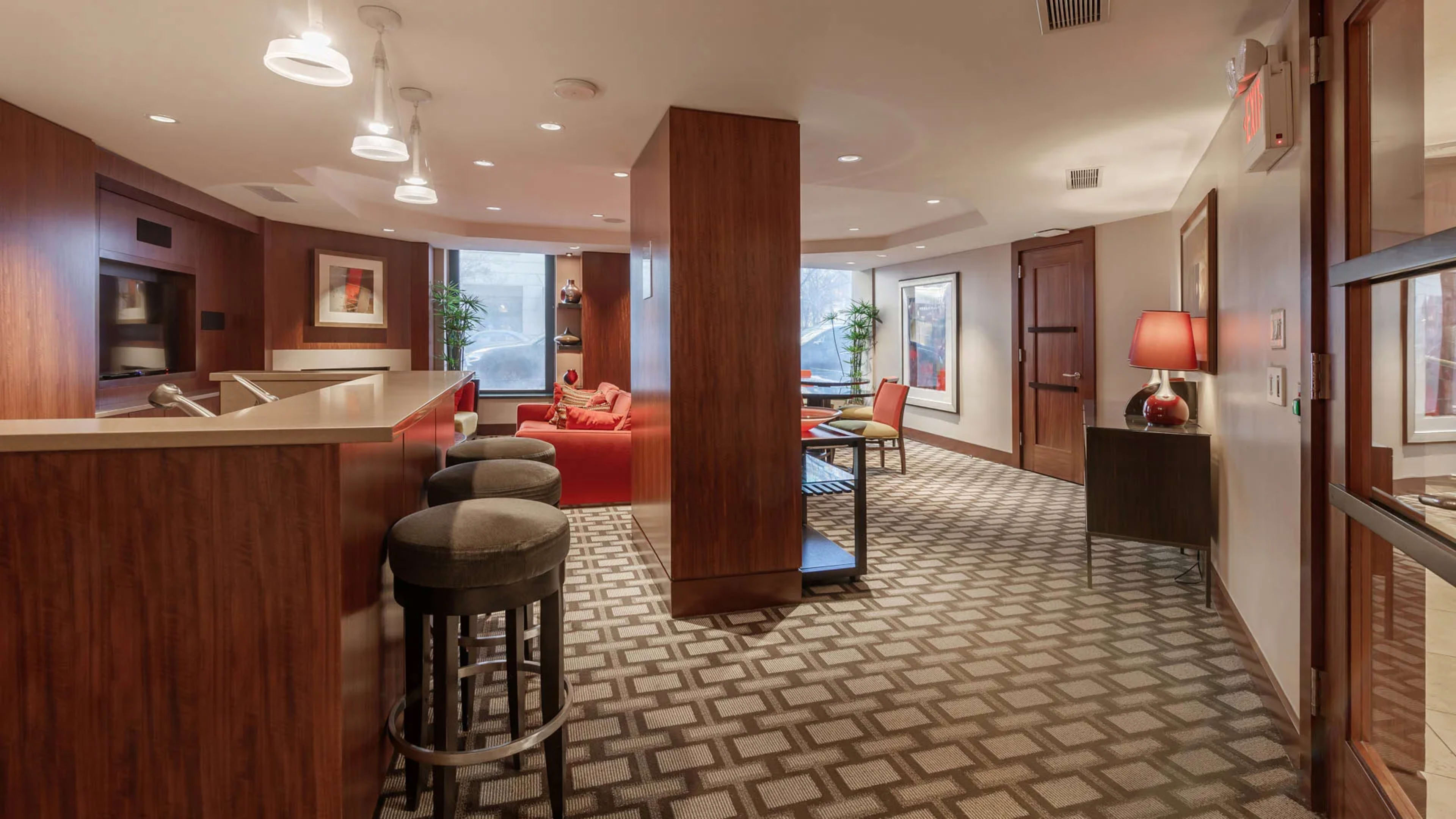 A modern lounge area with a bar, seating, and large windows, featuring wooden paneling and patterned carpet.