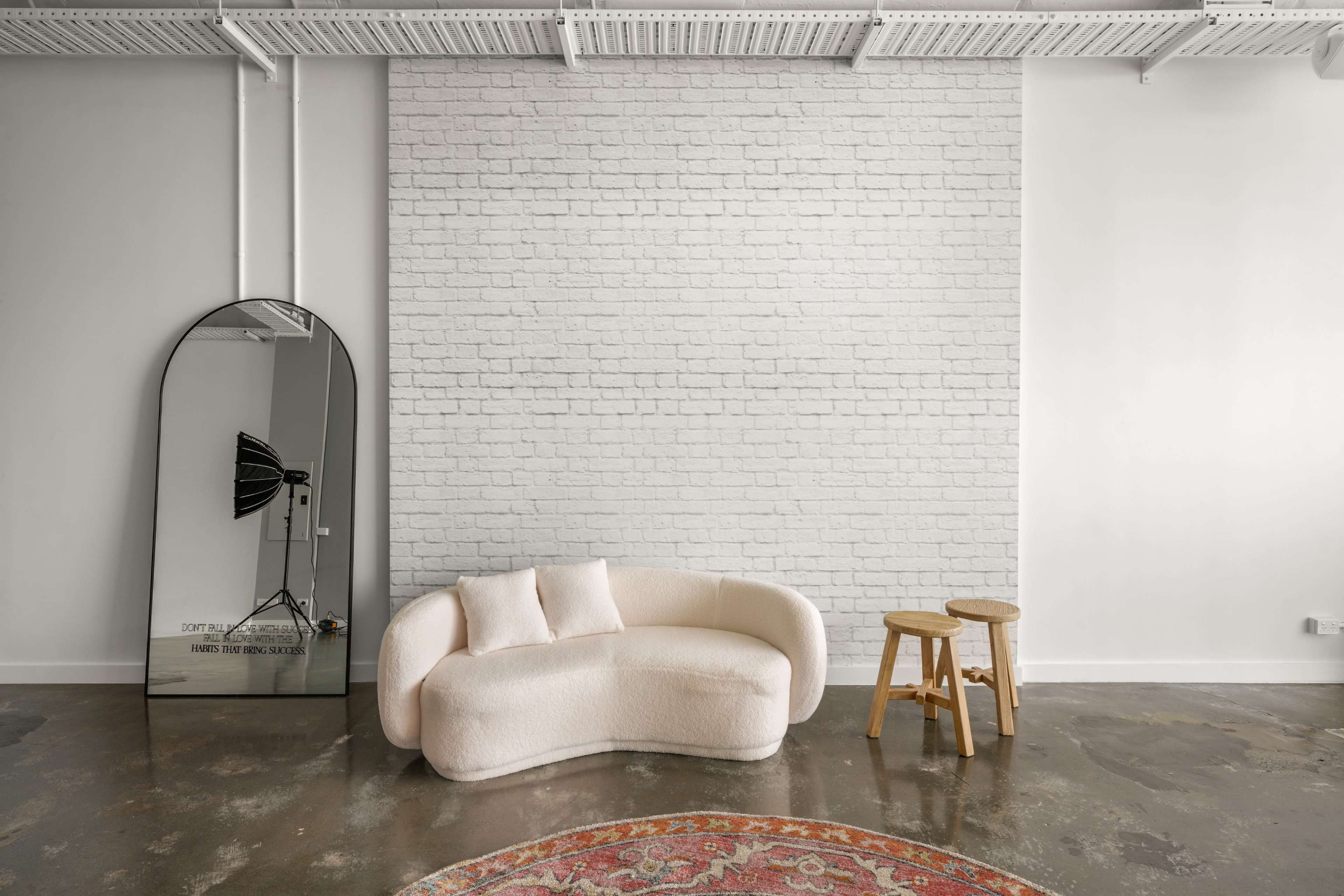 The image features a modern interior space with a white brick wall, a curved beige sofa, a wooden stool, and a large mirror reflecting light equipment.