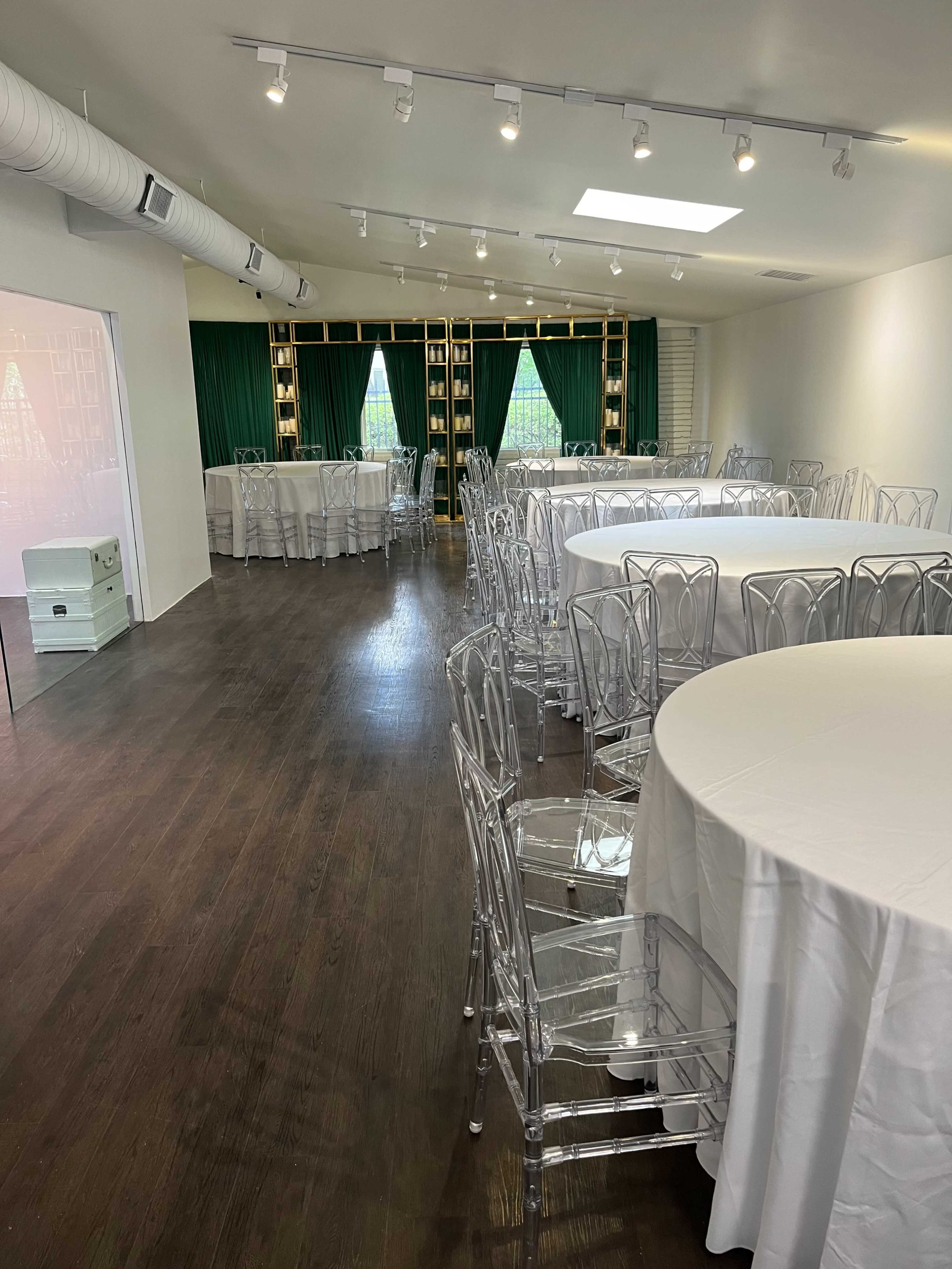 The image shows a spacious event room with circular tables covered in white tablecloths and transparent chairs arranged for a gathering, featuring green curtains and shelves in the background.