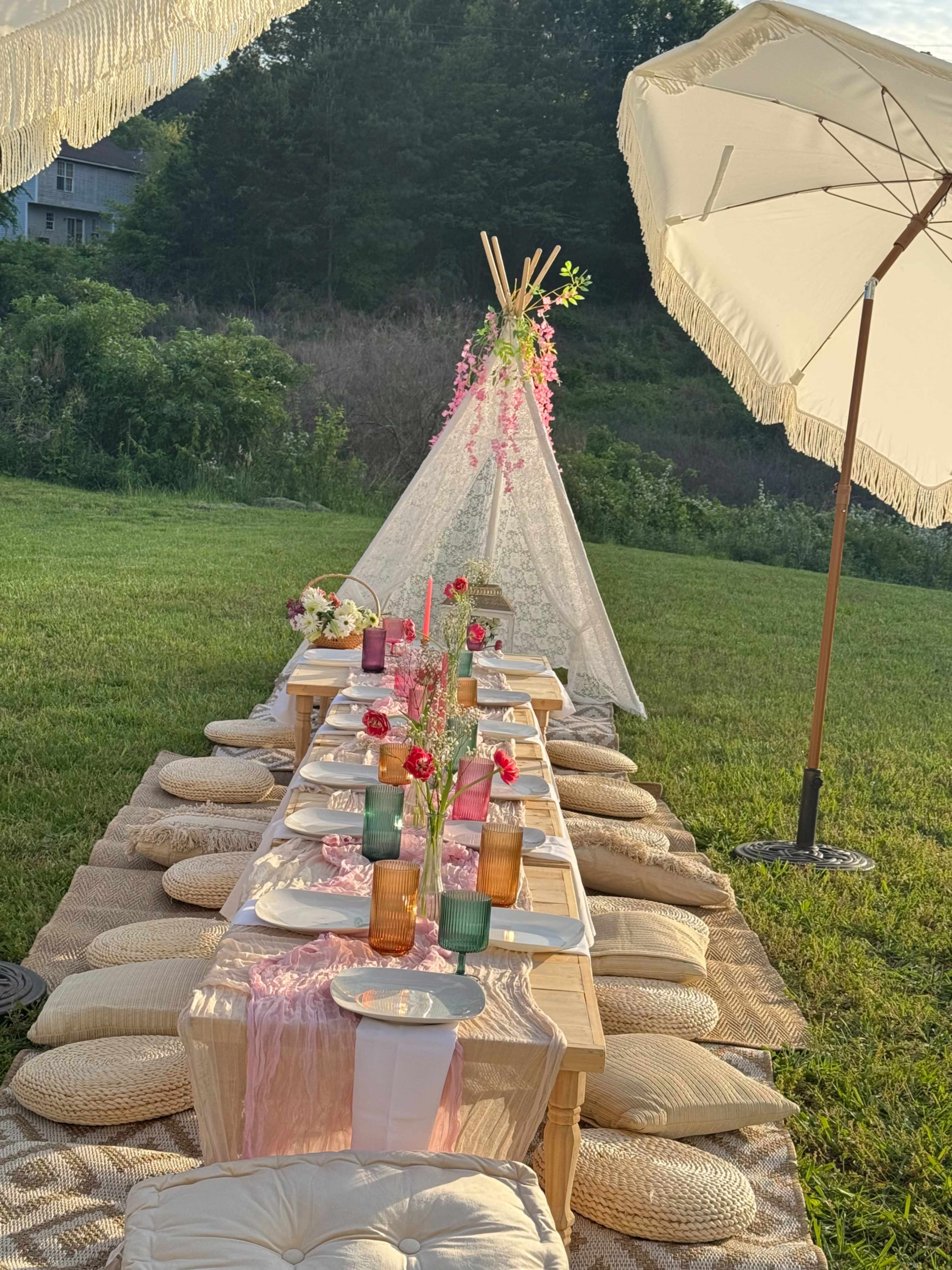 A long outdoor table is adorned with tableware, flowers, and candles, surrounded by cushions and shaded by large umbrellas.