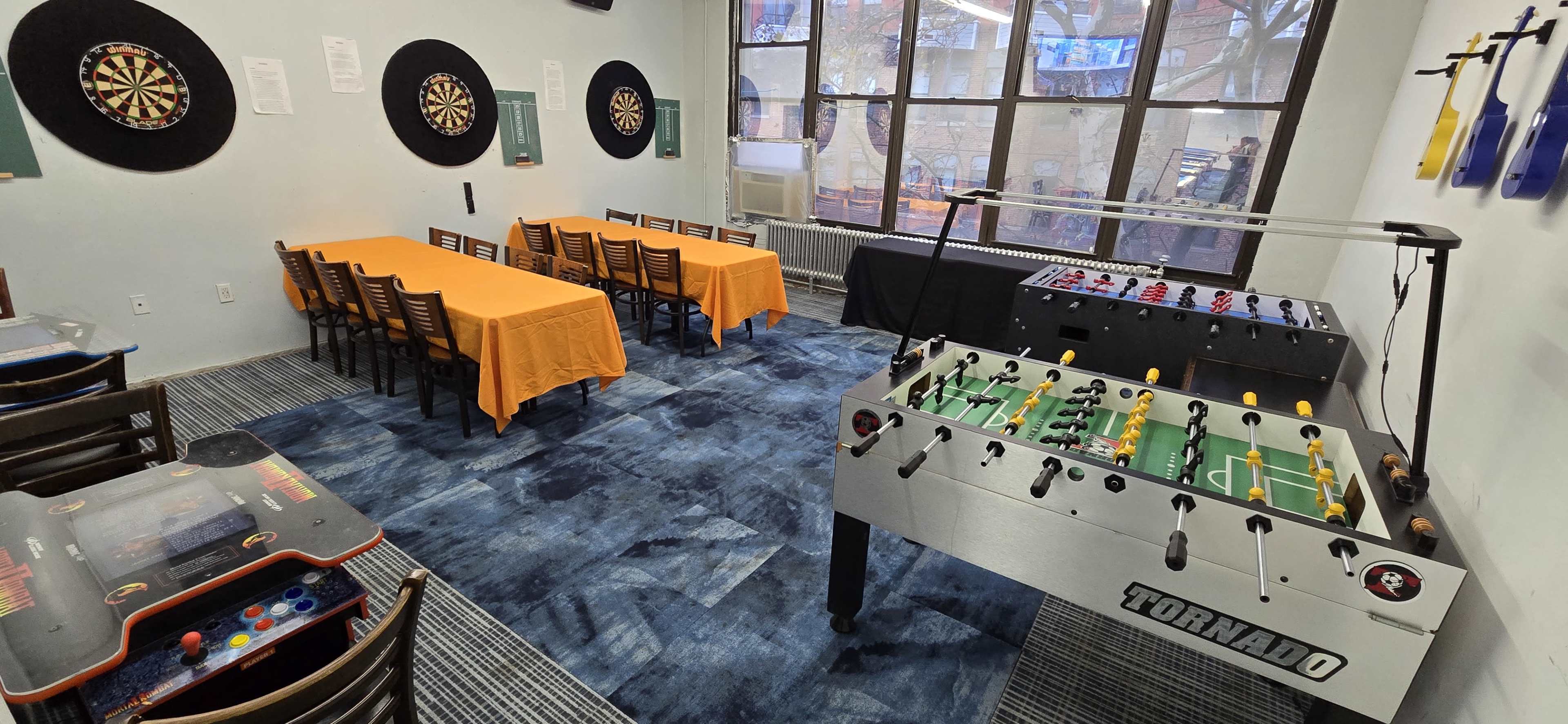 The image shows a recreational room with a table for dining, dart boards on the wall, and foosball tables.