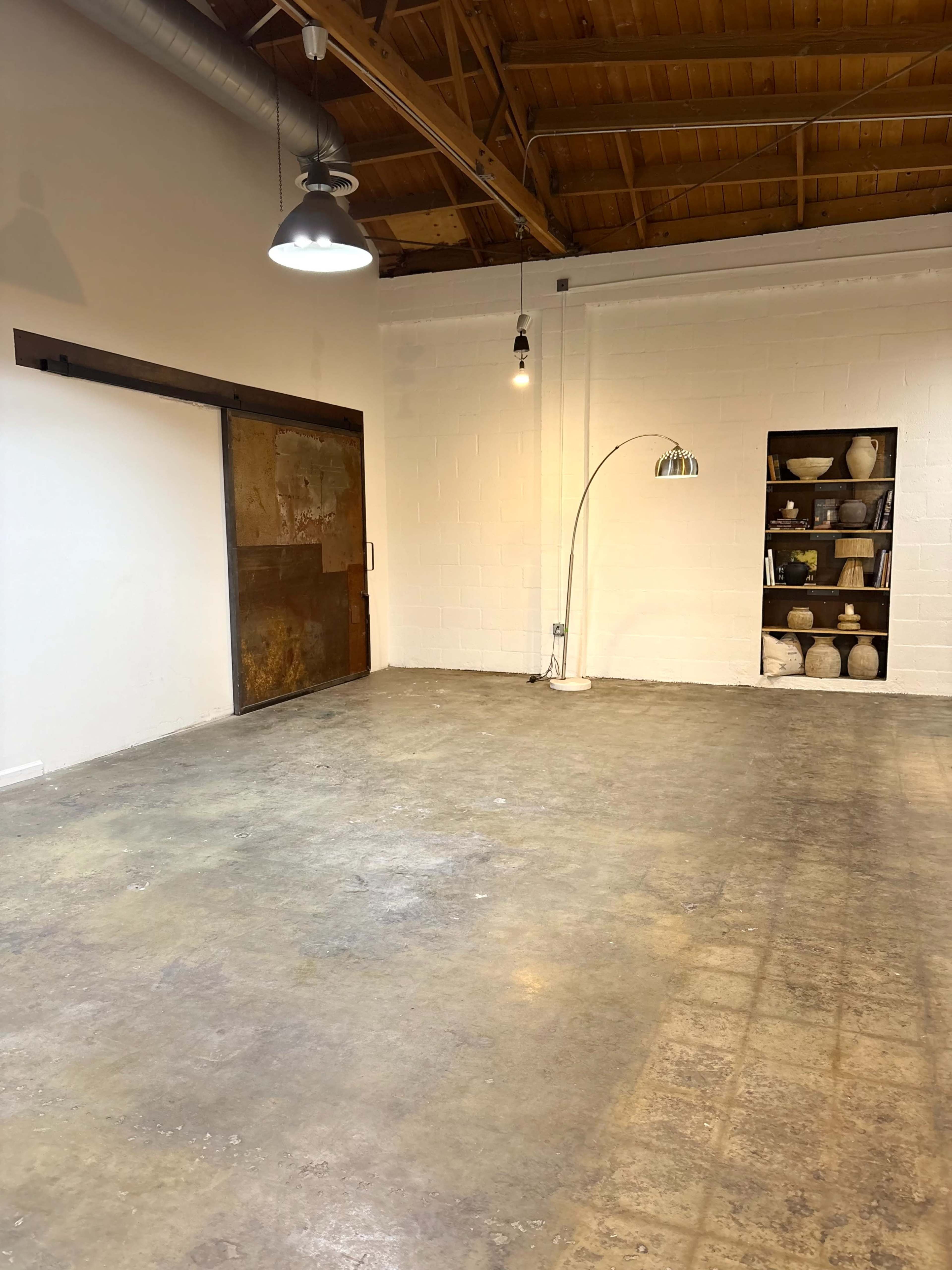 The image shows a spacious, empty room with a concrete floor, a large sliding door, a tall floor lamp, and a shelf displaying several pottery pieces.