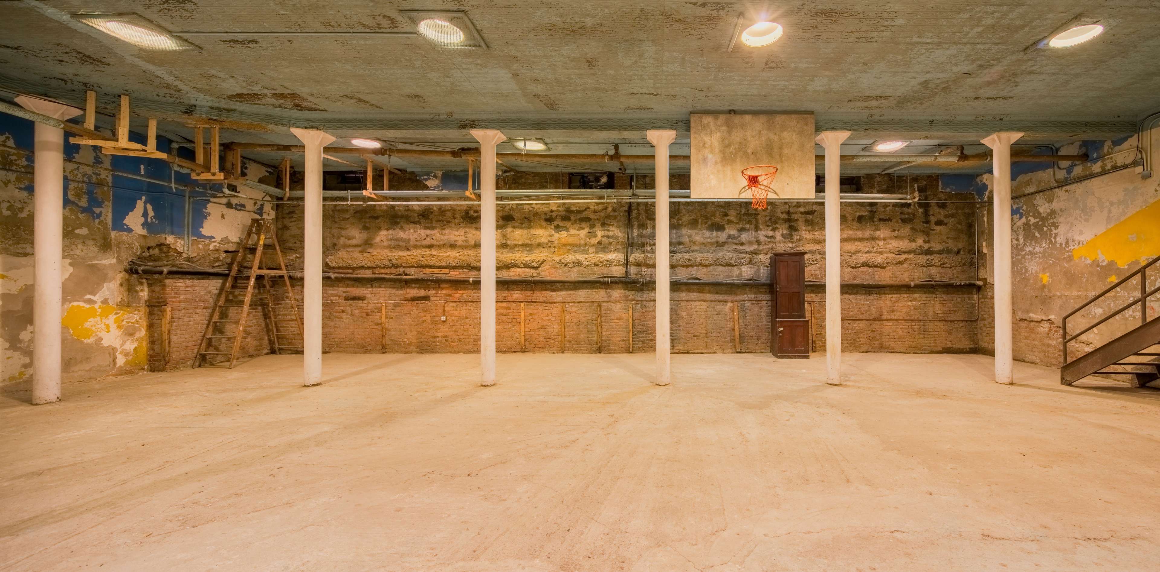 An empty underground basketball court with bare walls, exposed columns, and a faded basketball hoop.