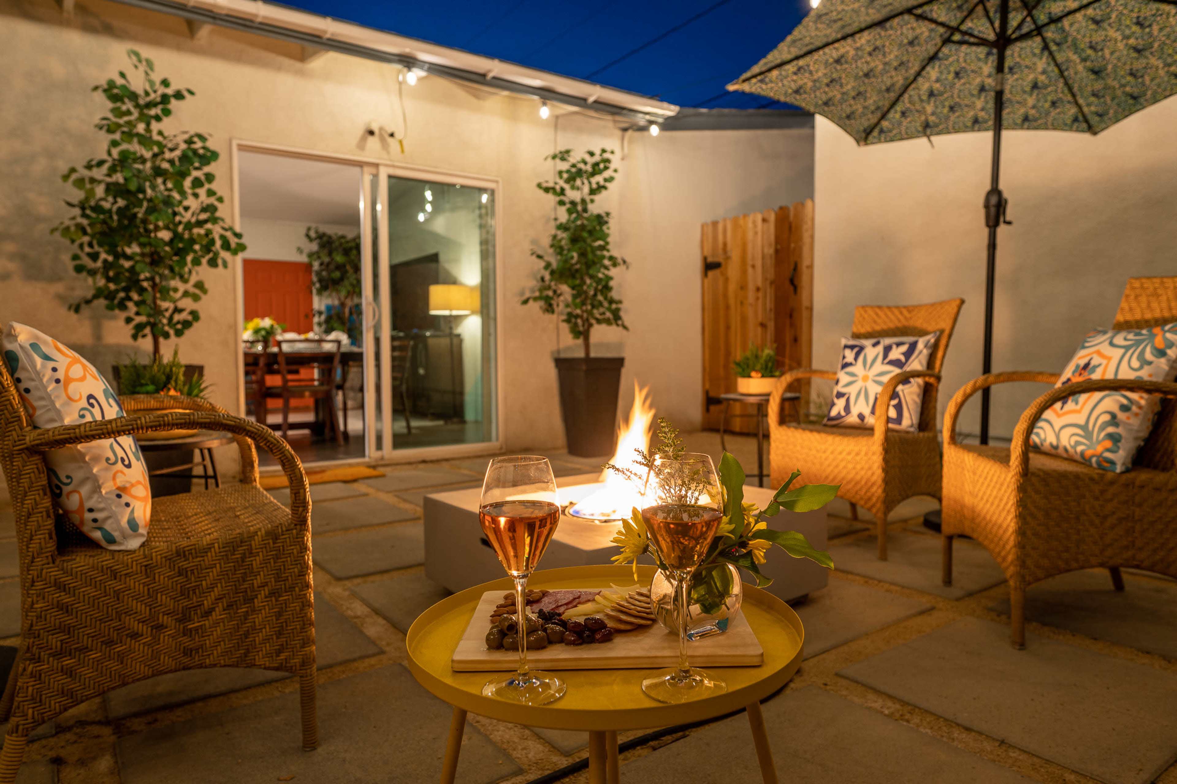 A cozy outdoor seating area features two glasses of rosé wine and a charcuterie board beside a fire pit, surrounded by chairs and umbrellas under a twilight sky.