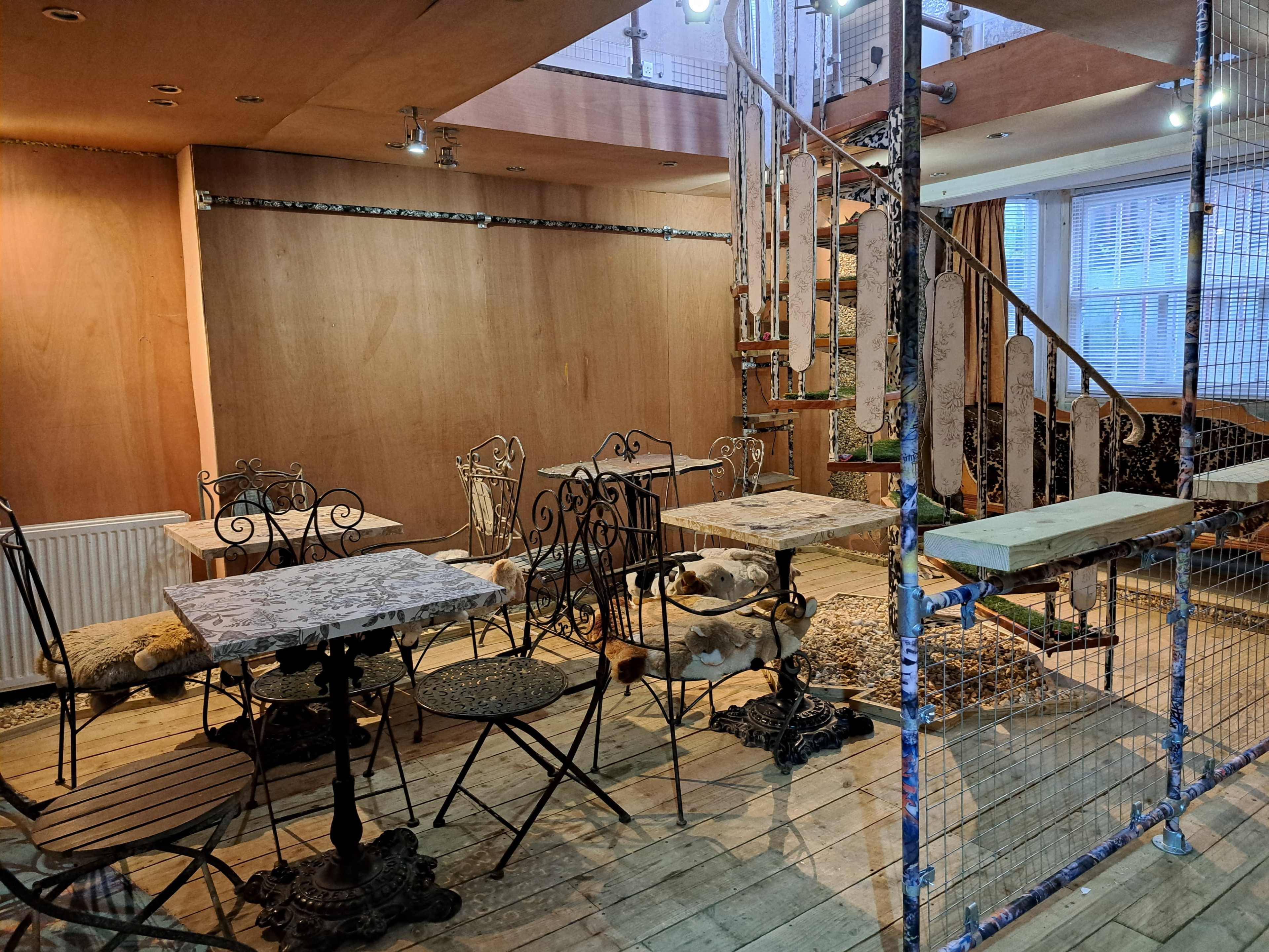 A café interior features metal tables and chairs arranged on wooden flooring, with a spiral staircase in the background and wooden paneling on the walls.